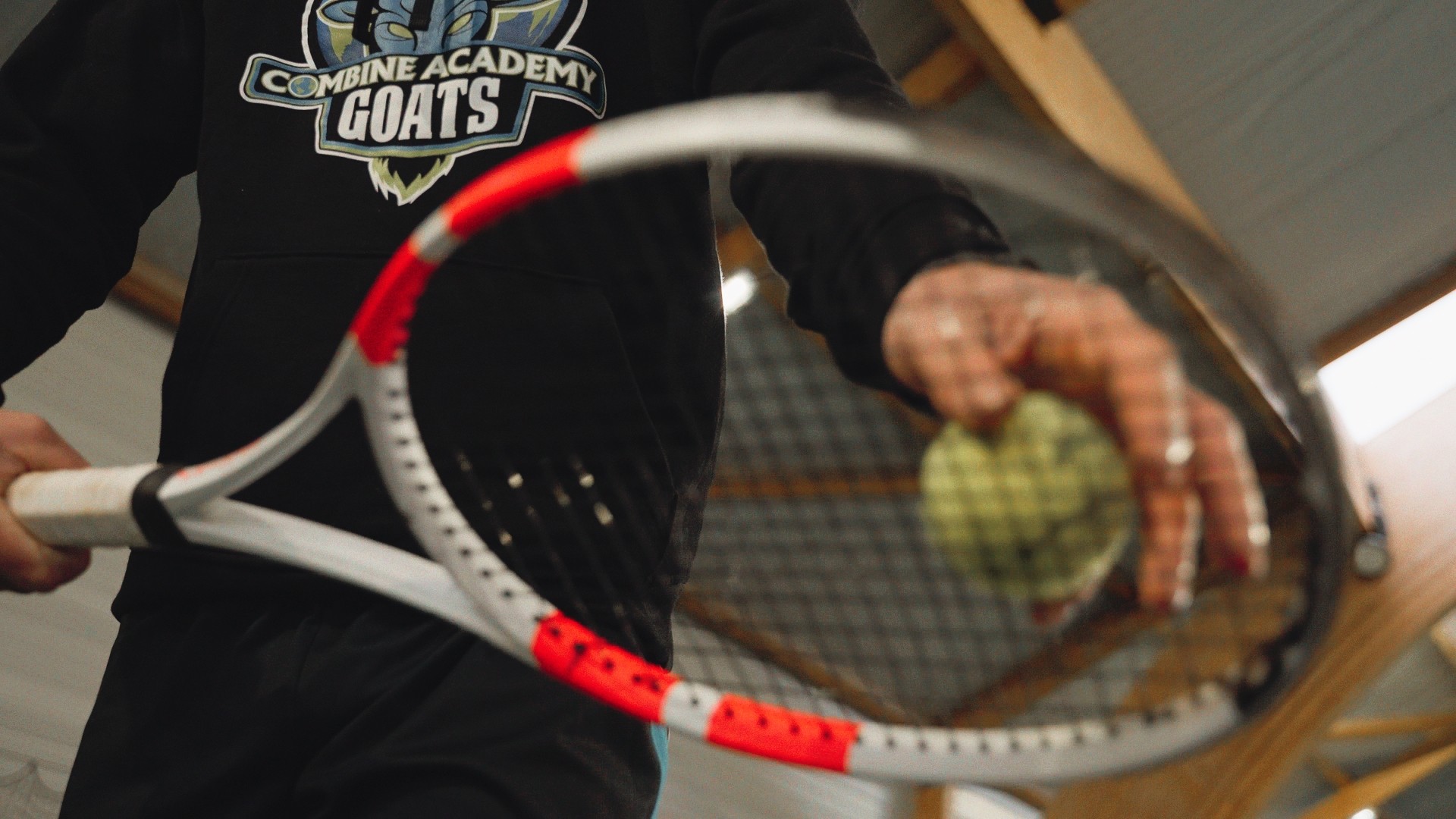 Gros plan d’une main plaçant une balle de tennis sur une raquette, sweat Combine Academy GOATS.