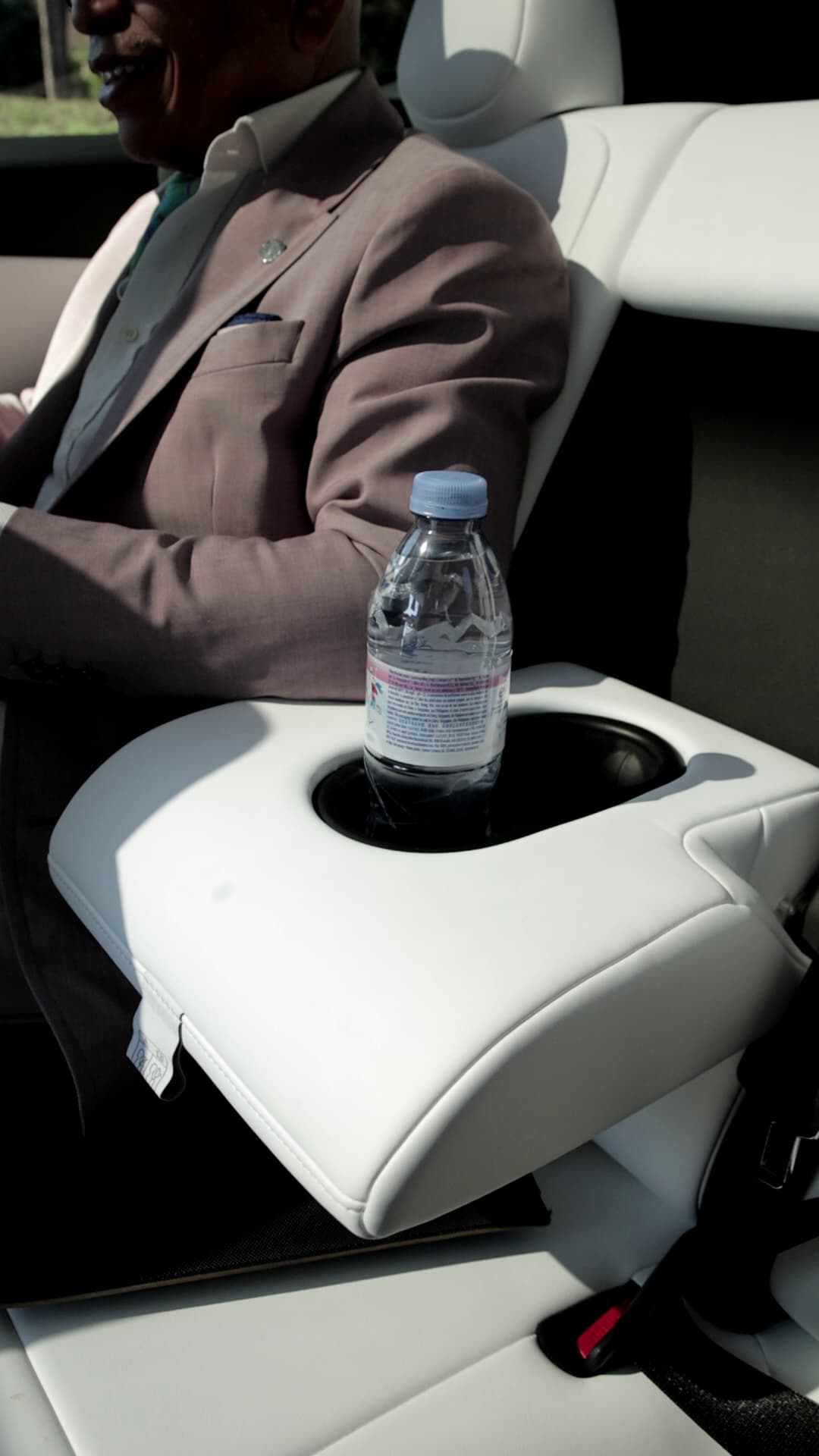 A focused shot of a water bottle on a Tesla Y water holder seat with a brown suited gentleman sitting beside it.