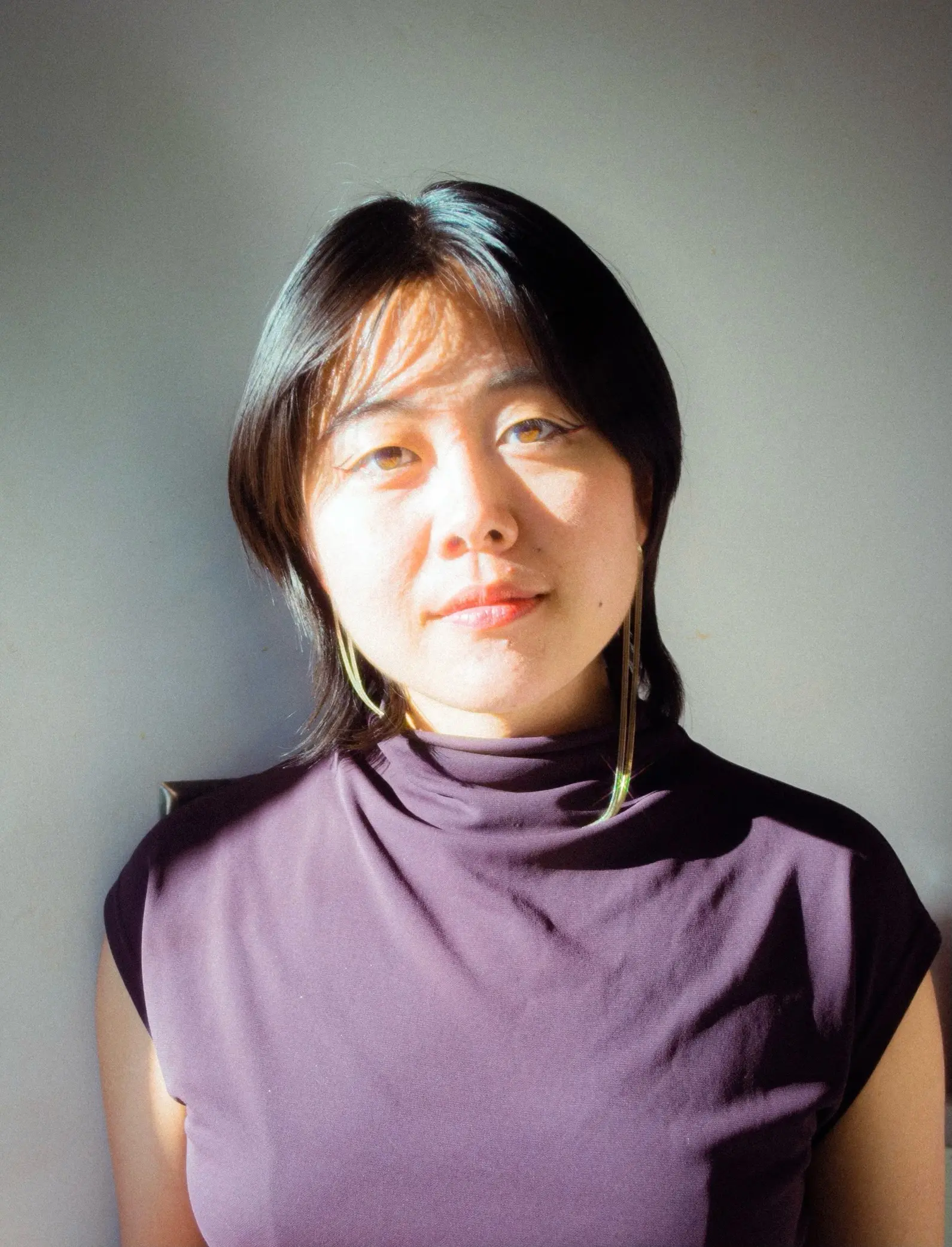 Kaori, with shoulder-length dark hair and dangling earrings stands against a plain light-colored background, wearing a dark, sleeveless turtleneck top, partially illuminated by sunlight.