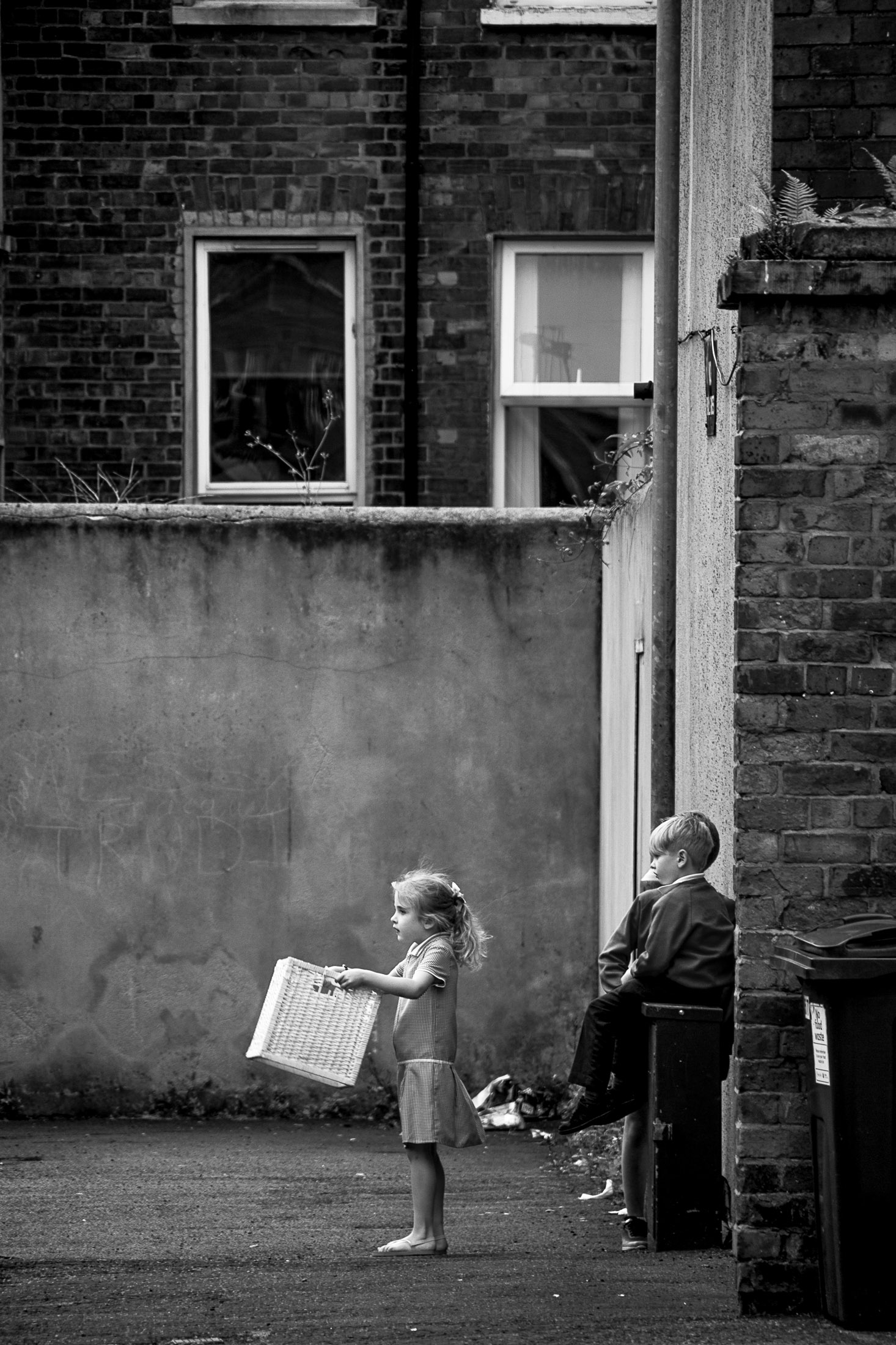 Two children standing in an alley behind brick buildings, documentary black and white street photograph.