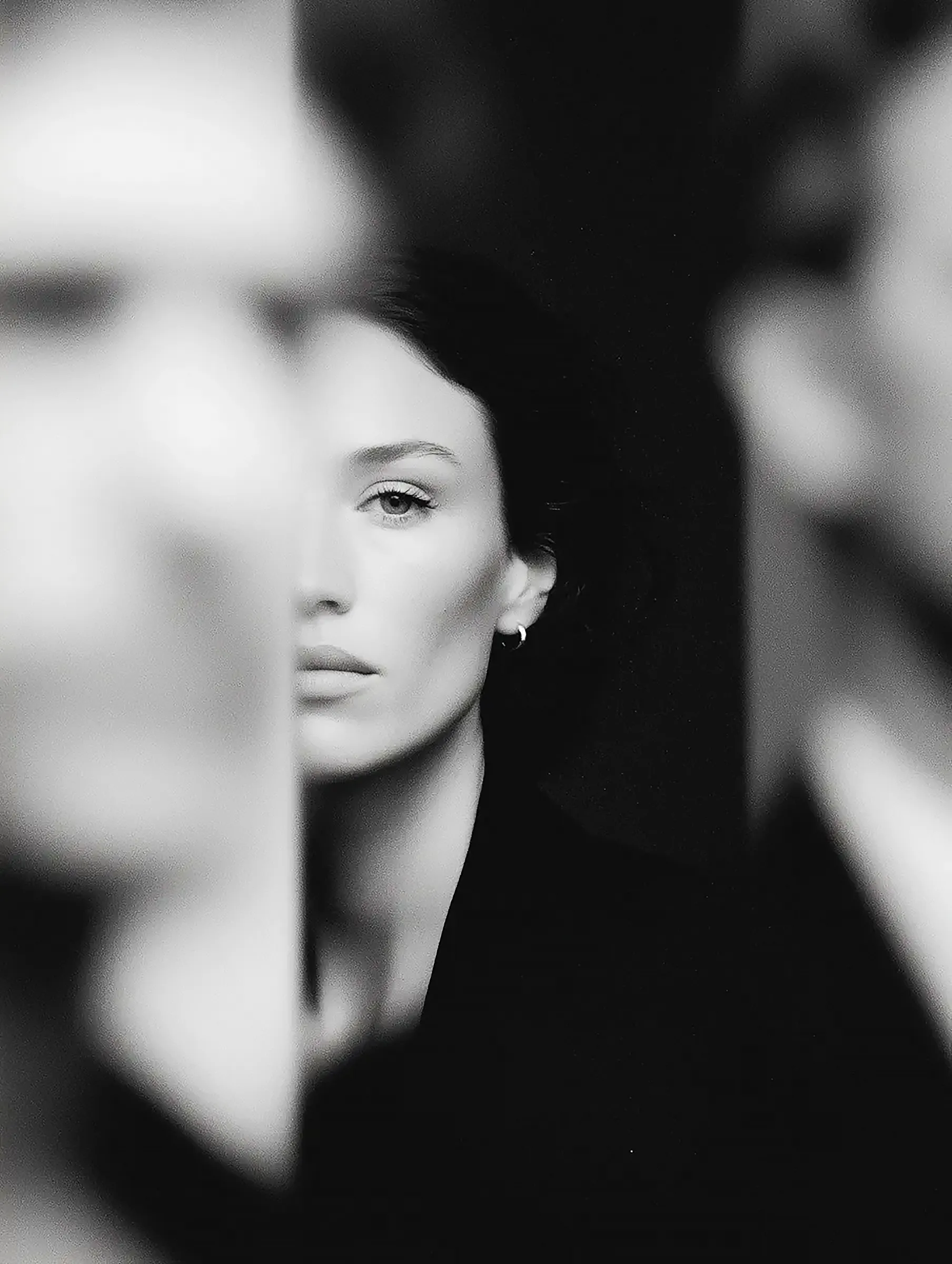 Black-and-white portrait of a woman with dark hair, partially obscured by blurred figures