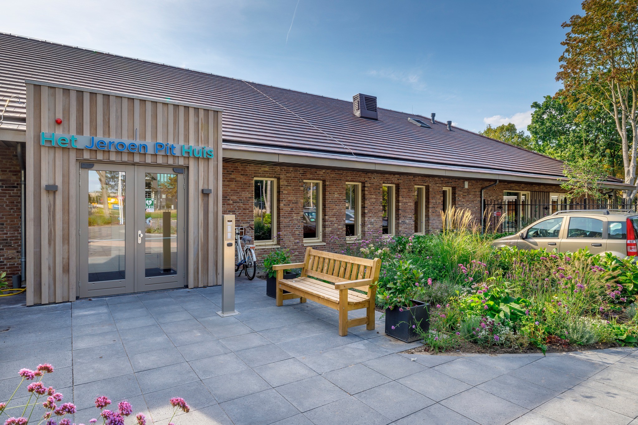 Het Jeroen Pit Huis, een bakstenen gebouw met houten details, bankje en bloemen.