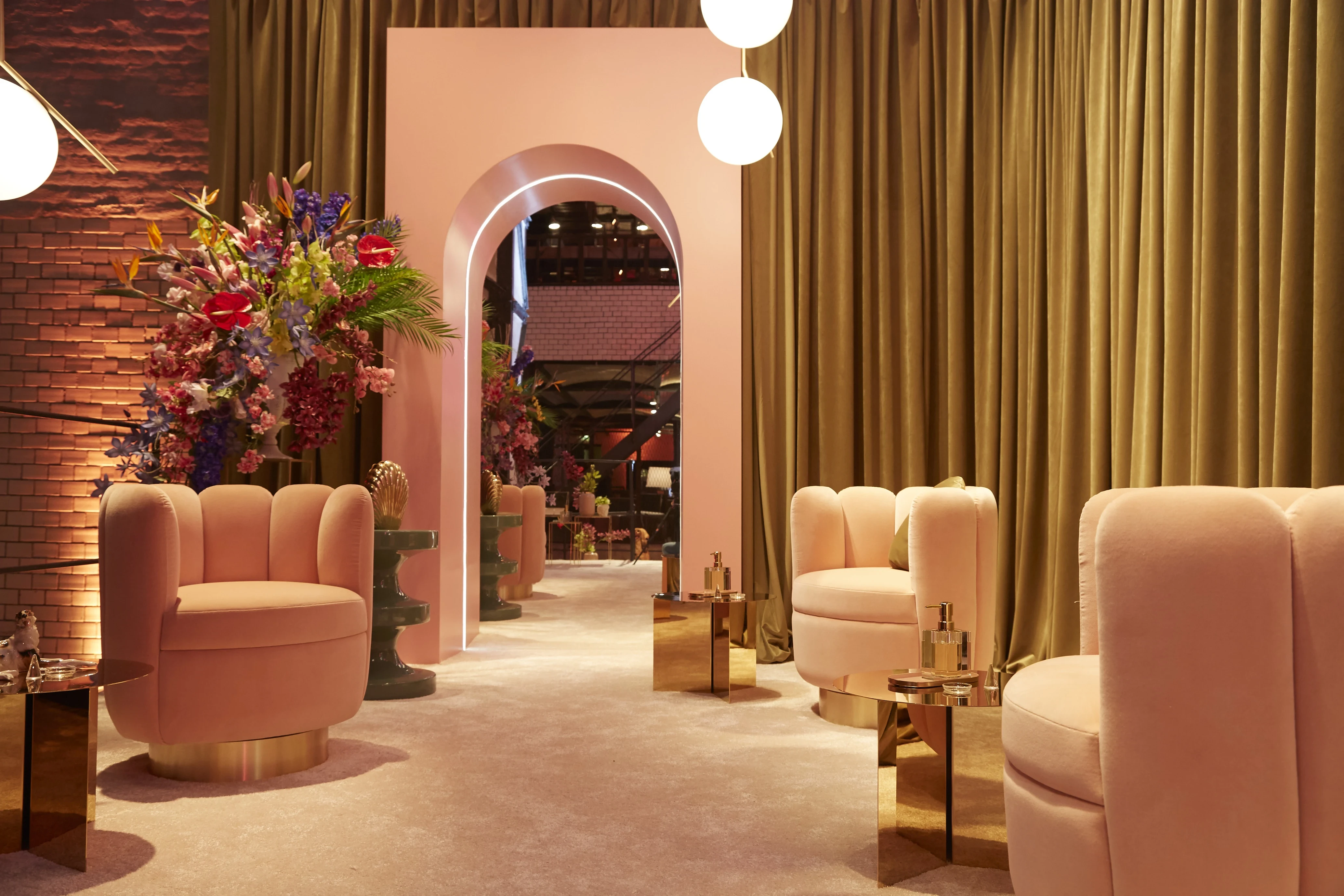 Blush-pink velvet armchairs around gold cube tables, framed by an arched portal and olive curtains at the Klarna Fashion Week lounge.