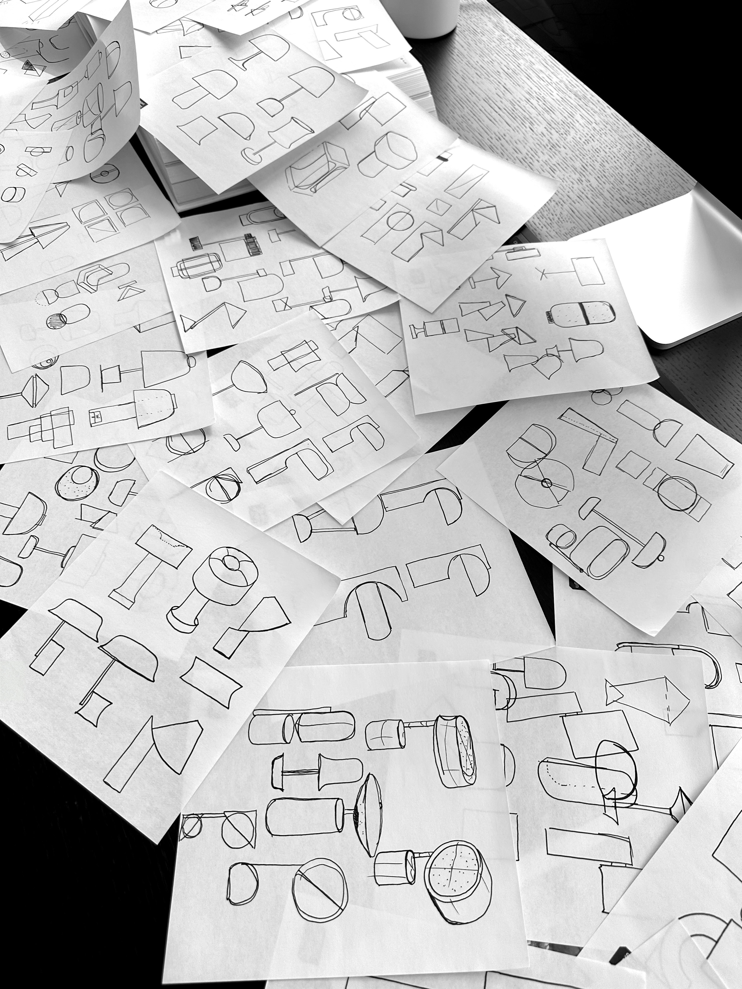 Pages of black ink sketches of lamp forms spread across a dark wood table