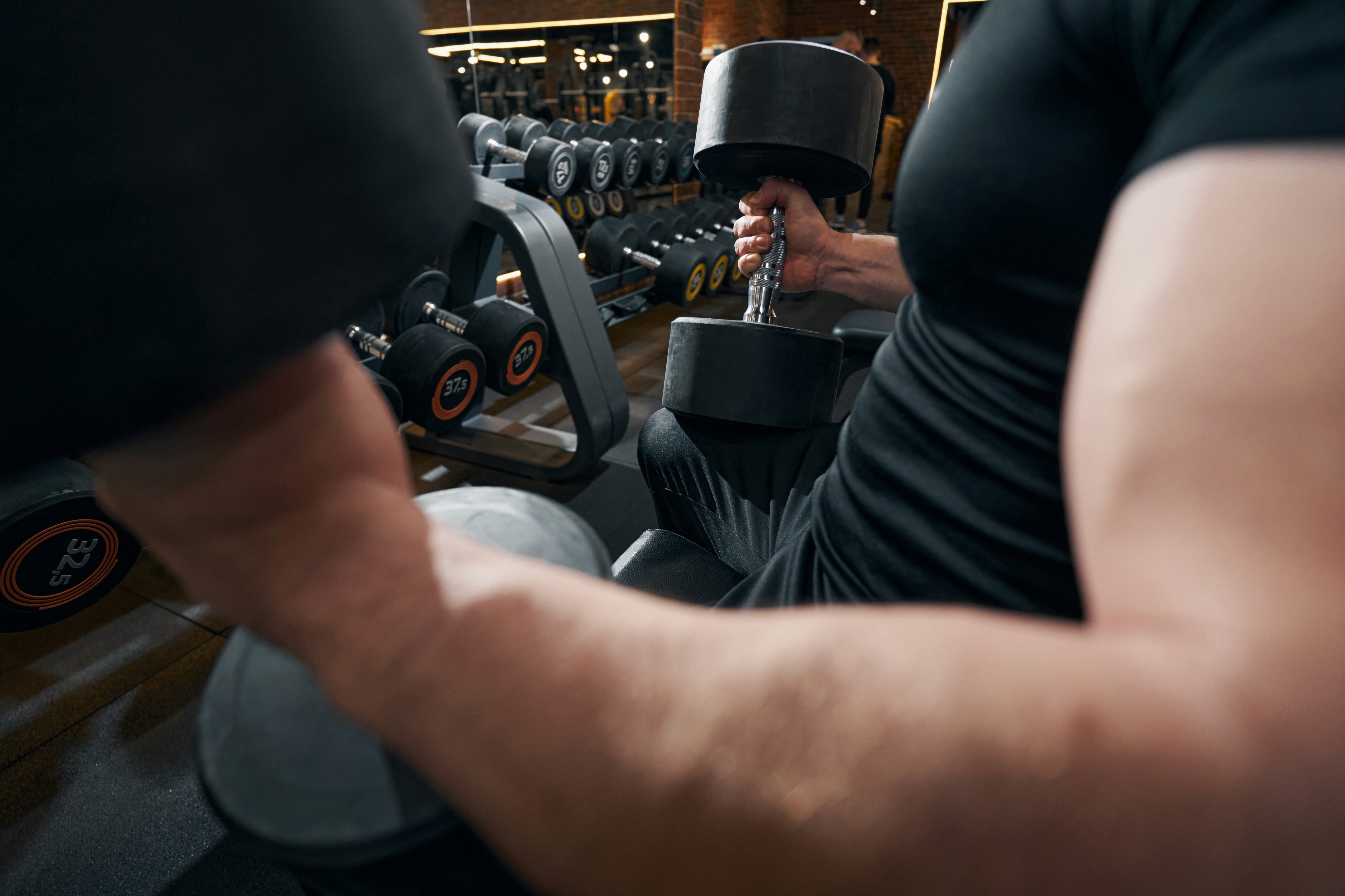 A person lifting a heavy dumbbell while seated in a gym. The focus is on the individual's muscular arms and the weight they are lifting