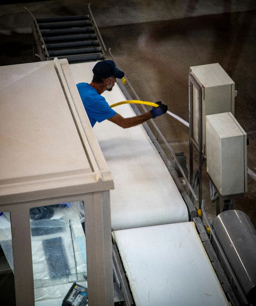 Ein Arbeiter mit blauer Kappe und blauem T-Shirt steht an einer Kontrollkabine und reinigt mit einem gelben Hochdruckschlauch eine weiße Förderband-Anlage in einer Industriehalle.