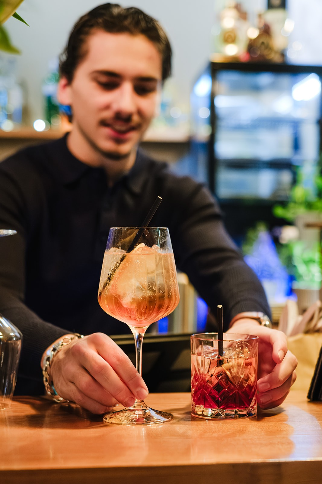 A bartender places two drinks on the counter: a tall spritz-style cocktail with ice and citrus in a stemmed glass, and a shorter amber-coloured drink in a patterned tumbler. The bartender smiles in the background as he serves the drinks.