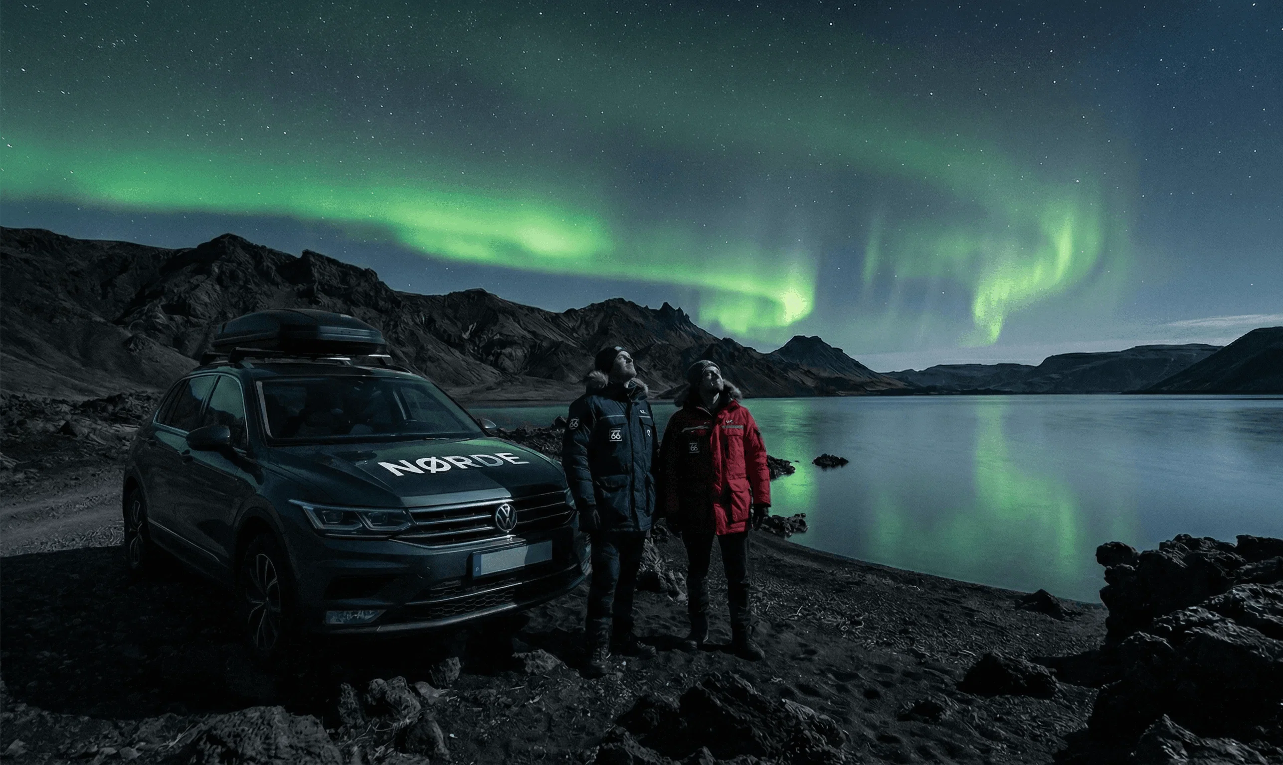 Two people standing next to a dark SUV parked by a lake at night under a strong display of the Northern Lights.