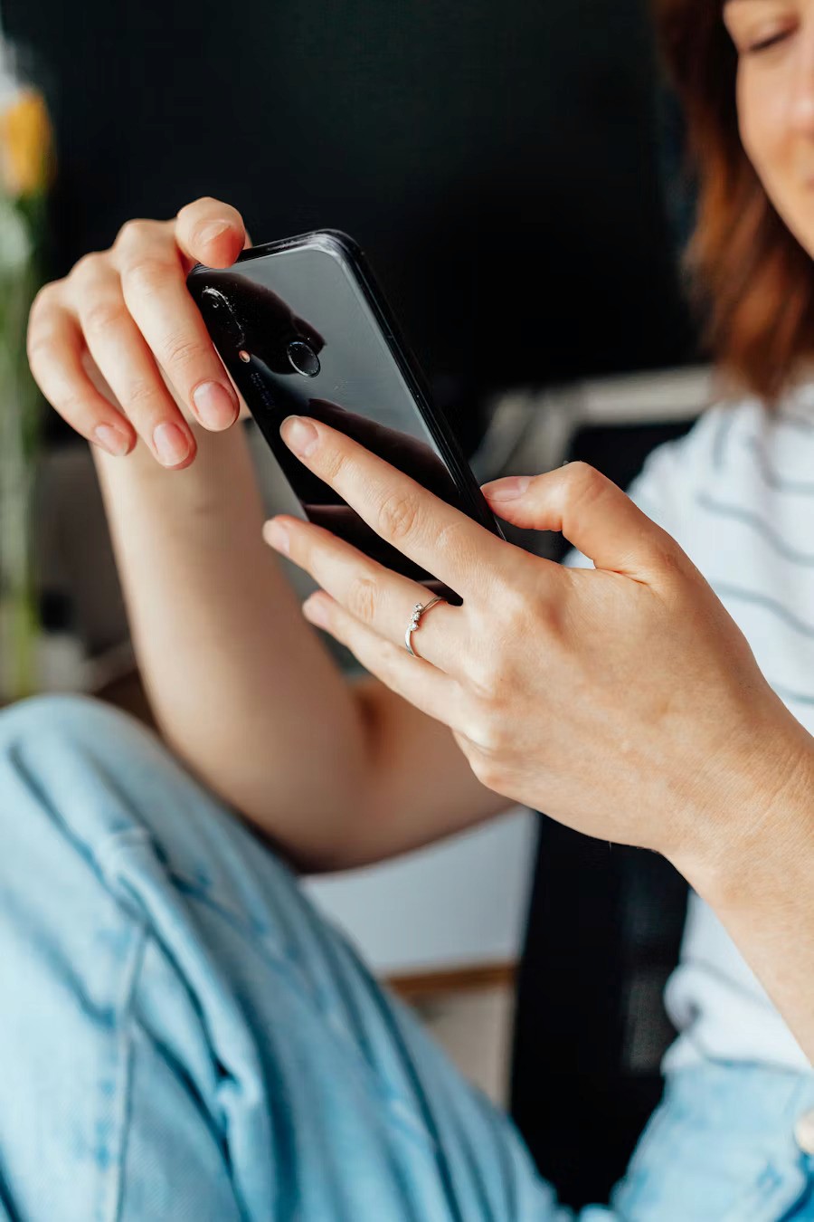 A person wearing a striped shirt and light blue jeans is holding a black smartphone, focusing on the screen, with a blurred background highlighting a vase with a single flower.