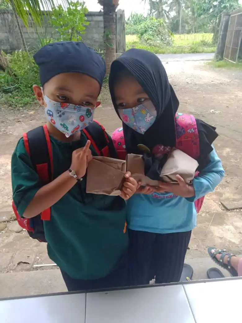  Two young Indonesian students in face masks and backpacks hold donated meal packs on their way to school