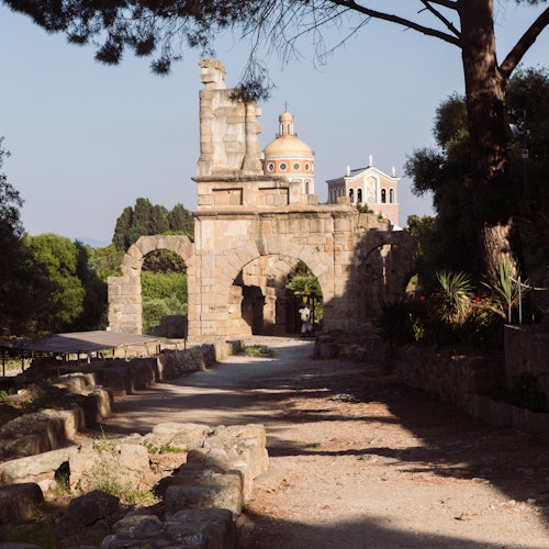 Arc antique en pierre avec un chemin qui le traverse. Deux bâtiments à coupole en arrière-plan, entourés d'arbres et de verdure.