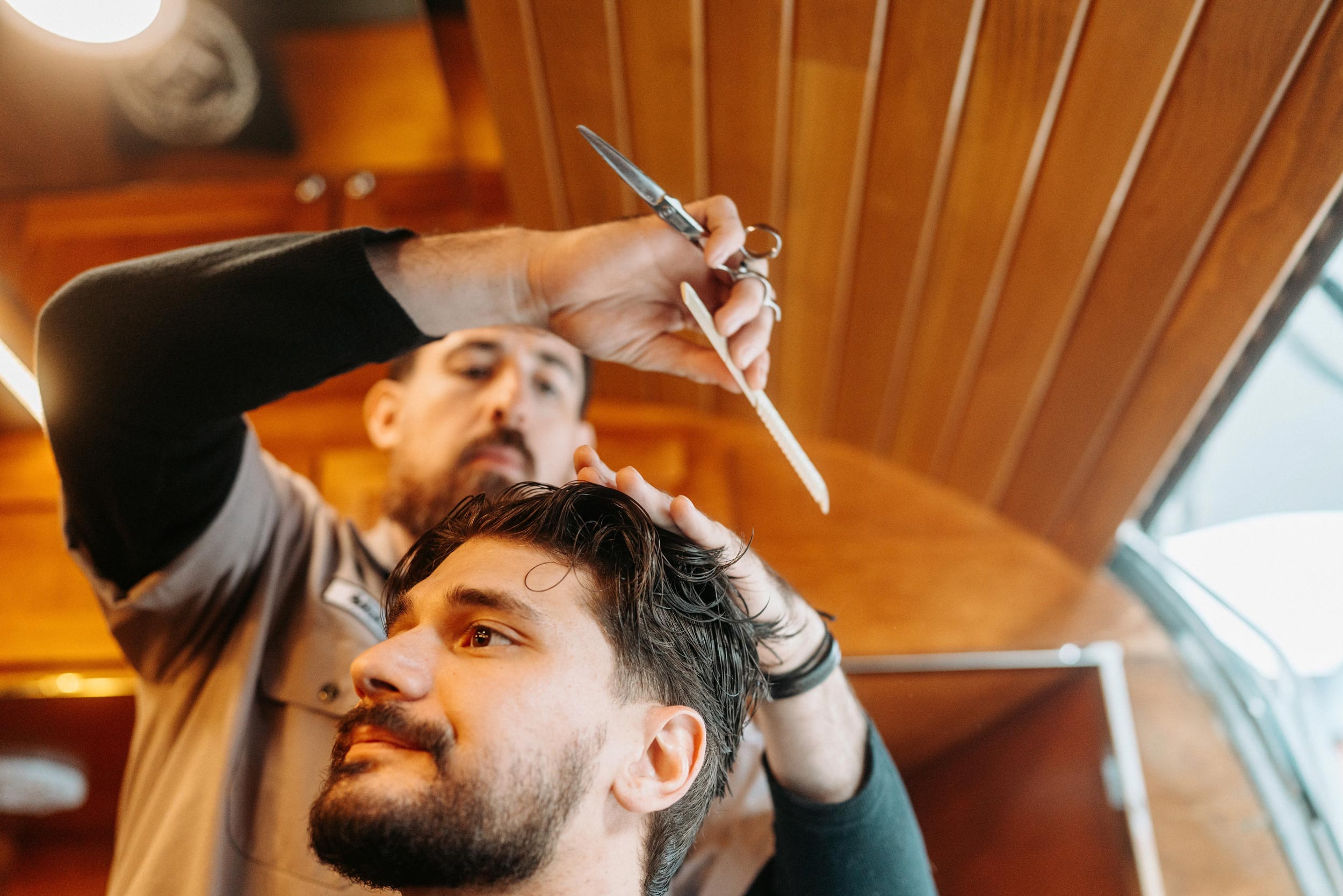 A close-up, eye-level shot of a bearded male barber, wearing a grey shirt, using a comb and scissors to cut the hair of a male client, who is looking up and away. The background is a warm, slatted wood paneling.