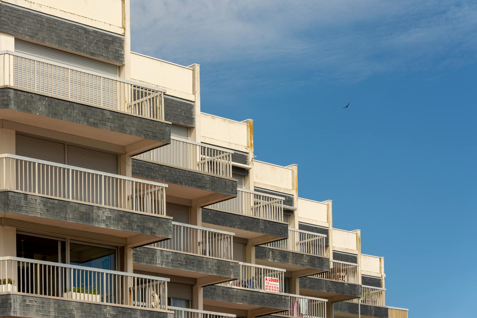 Modern apartment block with rows of white-railed balconies and grey-tiled panels against a clear blue sky with a small bird.