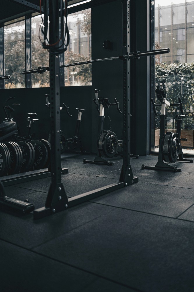 A dimly lit gym scene featuring a barbell rack and weights on the floor, with large windows in the background.