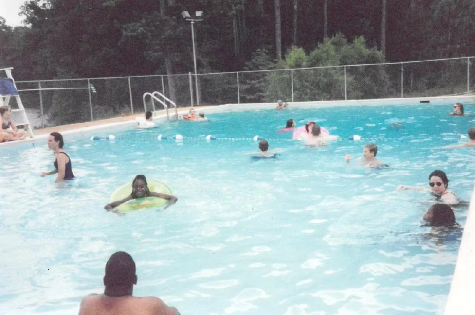 People of all ages enjoy a sunny day swimming in a pool. Green trees line the background.