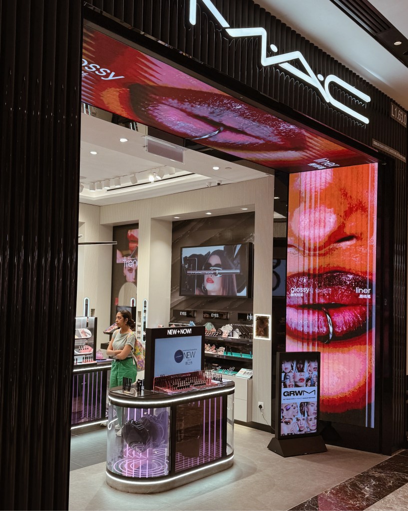 Interior view of a M.A.C cosmetics store featuring bold advertisements for lip products and a customer browsing at the counter.