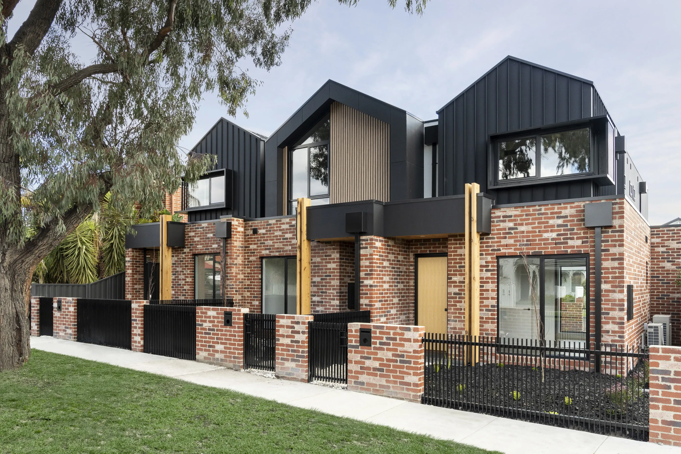 Street view of the Coburg townhouses, a row of homes with recycled-brick lower levels, black metal-clad upper gables, timber-batten accents, timber posts and a black picket and recycled-brick fence under a gum tree