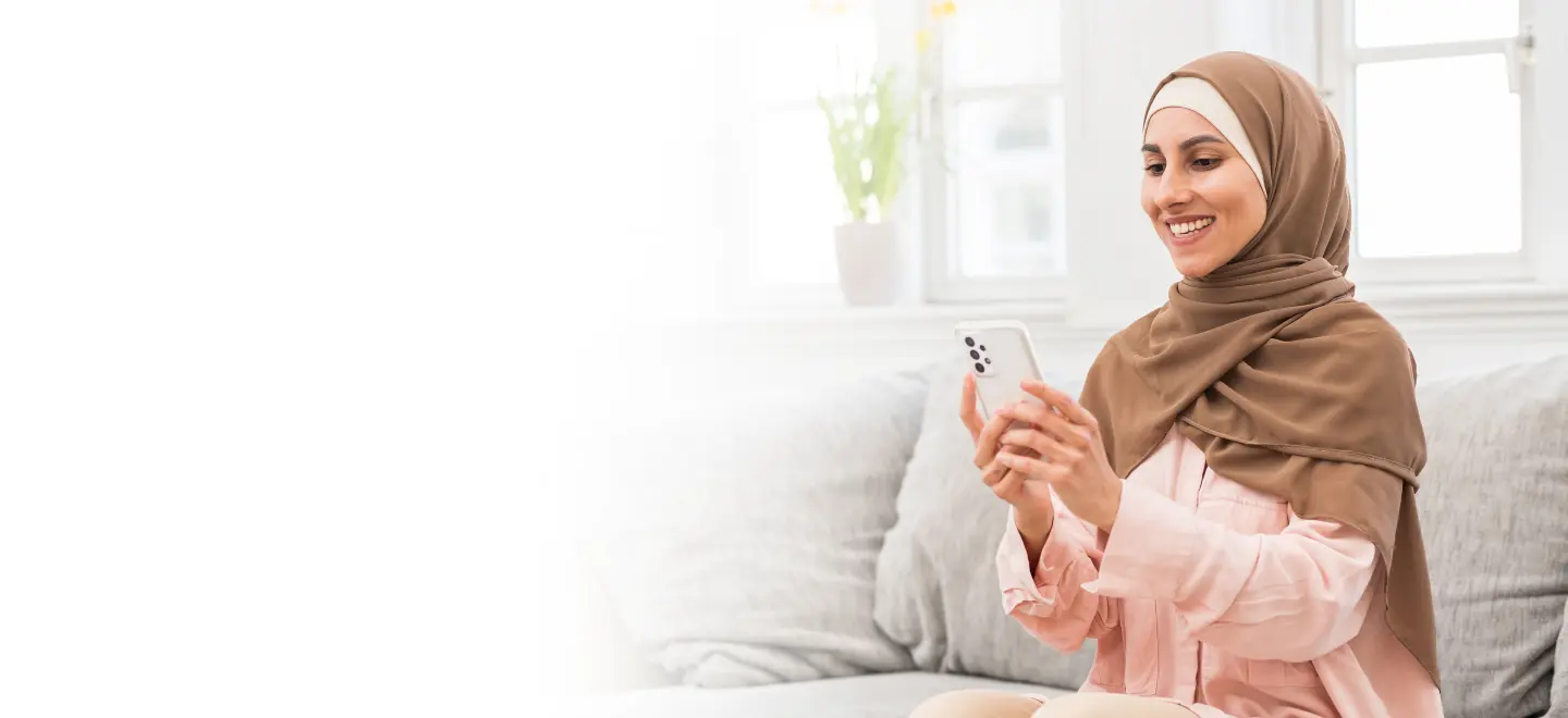 A smiling woman wearing a hijab sits on a couch while using a health app on her smartphone, showcasing digital health solutions for chronic conditions like diabetes in a comfortable, home setting.