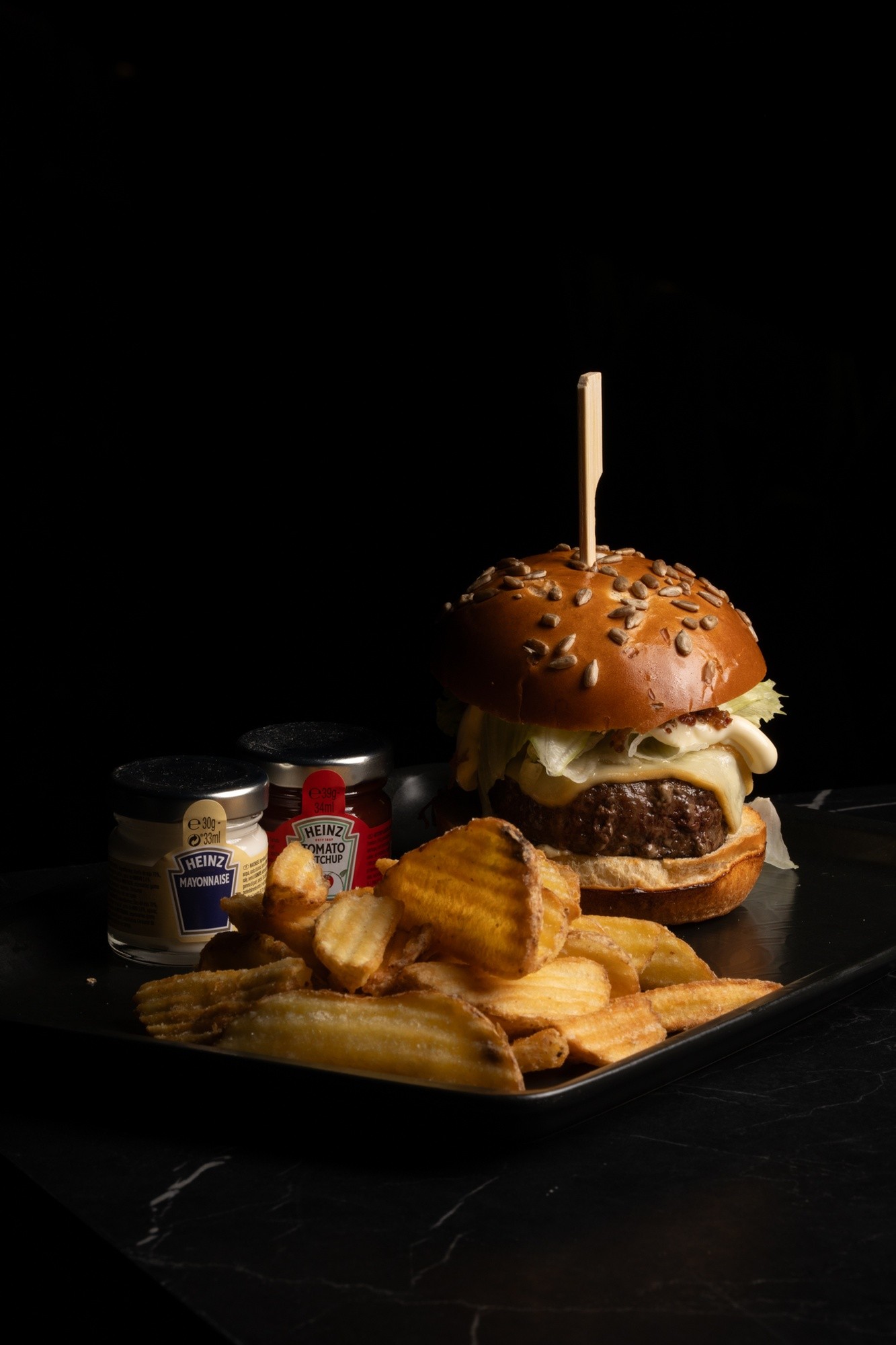 Hamburger di manzo con pane ai semi in superficie, bacon e lattuga su vassoio con patatine e barattolini di salse su sfondo nero