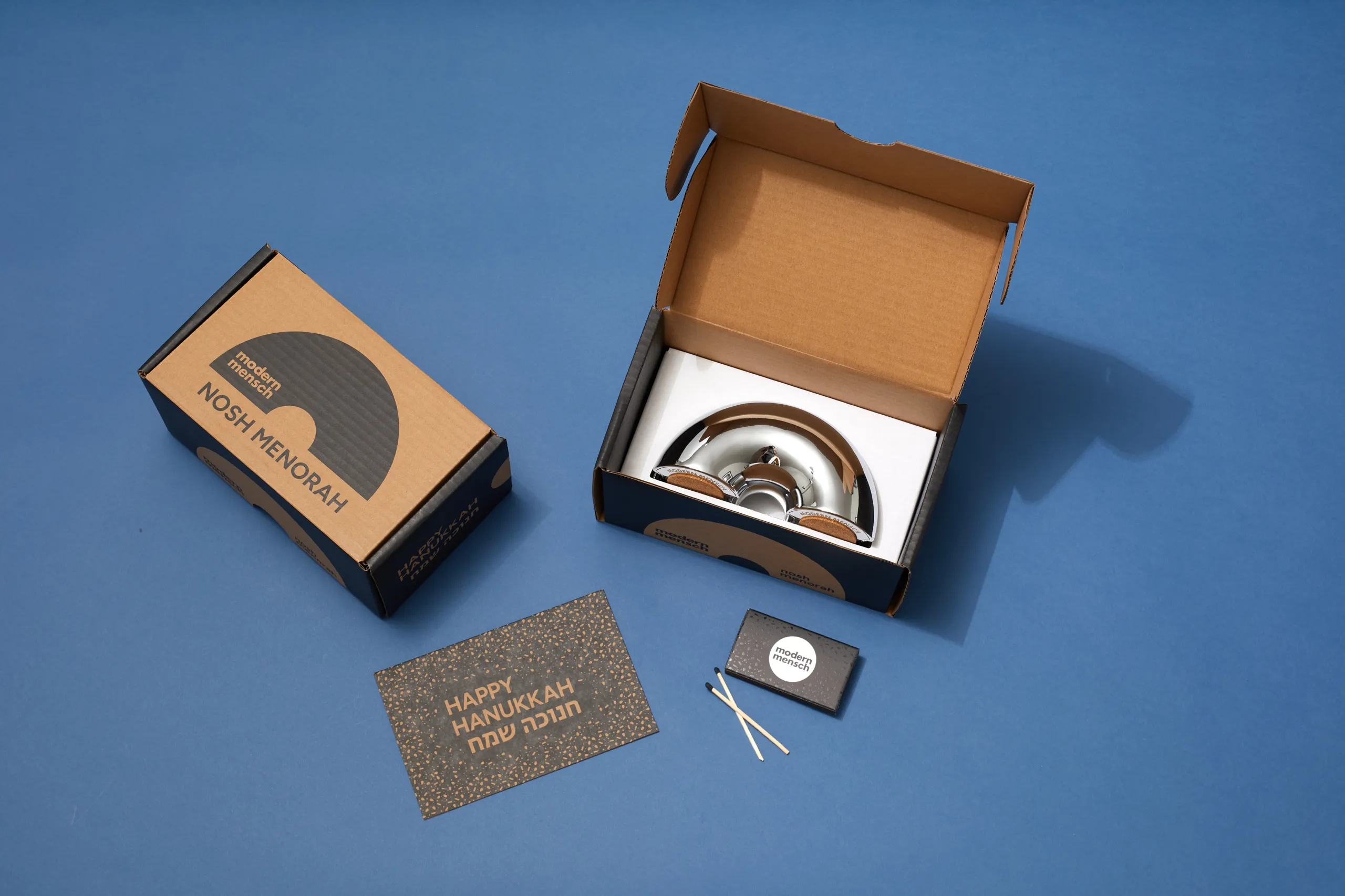 Open Modern Mensch Nosh Menorah packaging with menorah, card, and accessories displayed on blue
