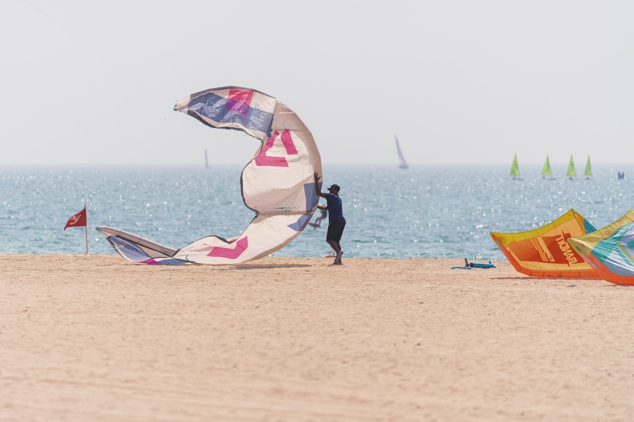 A kitesurfer is setting up a kite on a sandy beach with the sea and sailboats in the background.&nbsp;