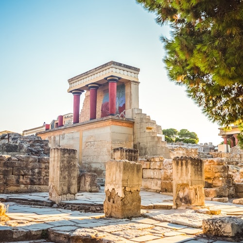 Ancient ruins with columns and a partially restored building featuring red pillars and fresco remnants under a clear blue sky.