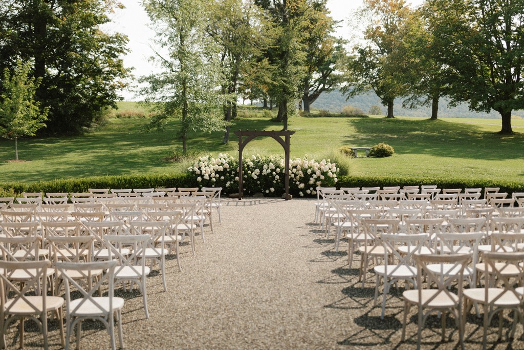 Garden wedding ceremony setup with white crossback chairs, a wooden arbor, and blooming white hydrangeas on a sunny estate lawn