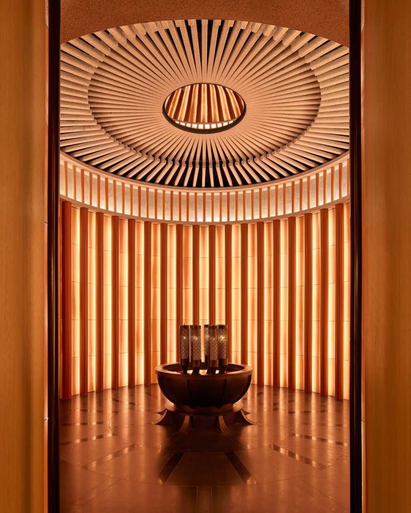 A warm, inviting interior of a spa or wellness center showcasing a circular ceiling with radial patterns, illuminated by ambient lighting. The walls are lined with vertical wooden panels, and a stylish bowl-shaped feature sits in the center, surrounded by decorative light fixtures.
