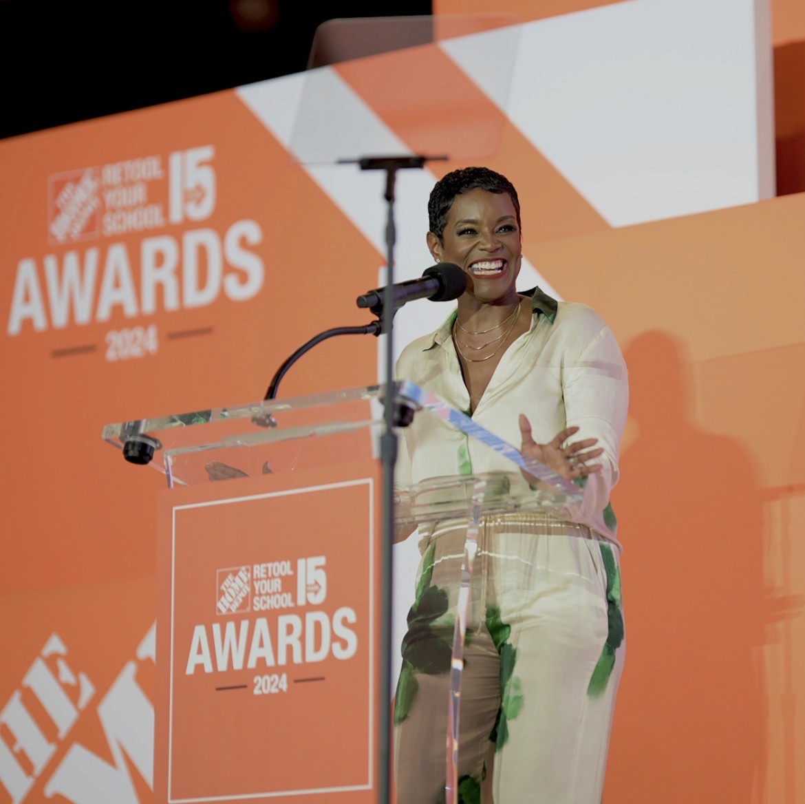 Rashan Ali Smith in a cream and green outfit speaks at a clear podium with a microphone at the 2024 Home Depot Retool Your School Awards. The stage background and signage are orange and white.