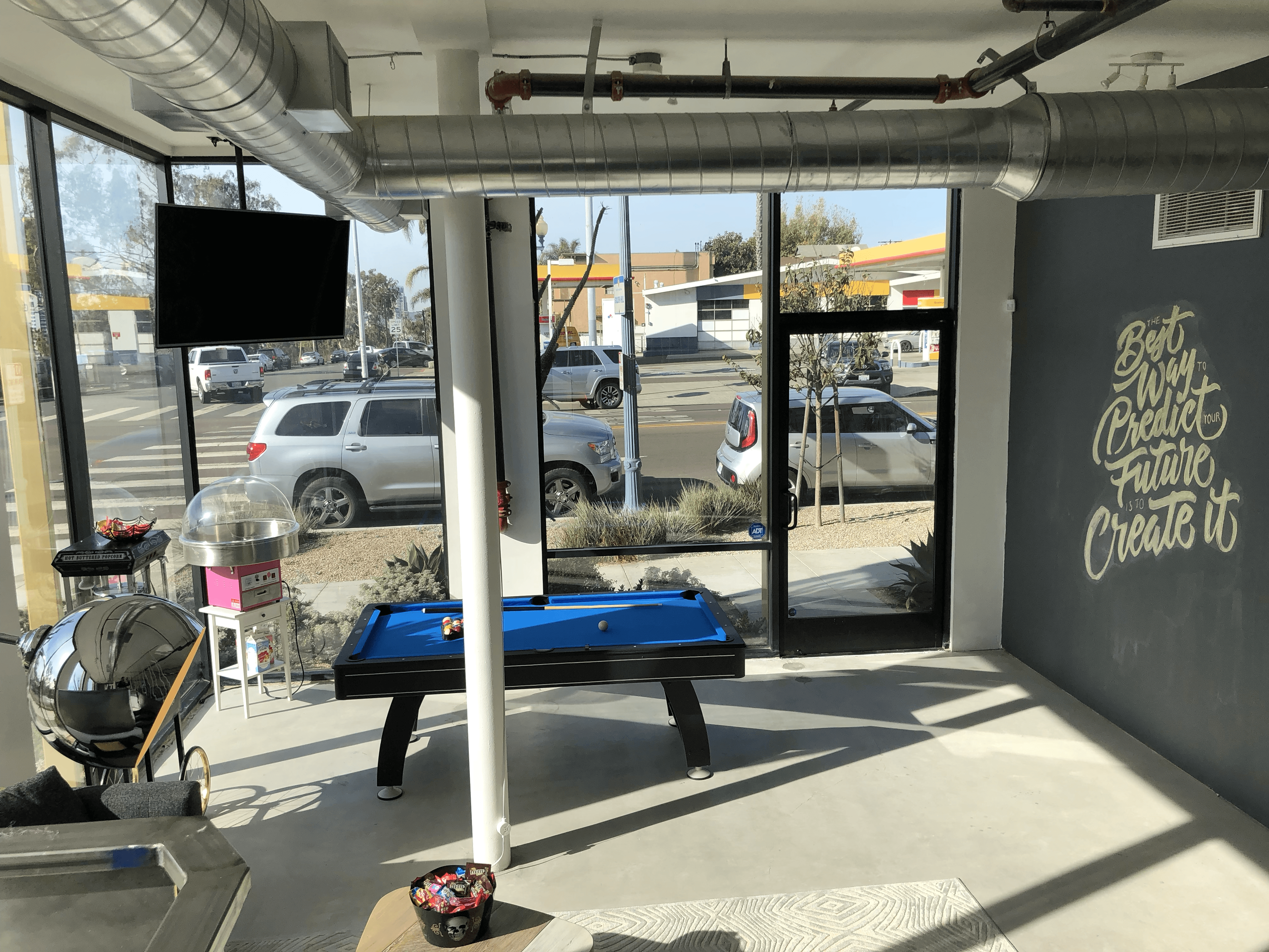 Interior of a gym with a blue bench, workout equipment, and a large window showing parked cars outside.