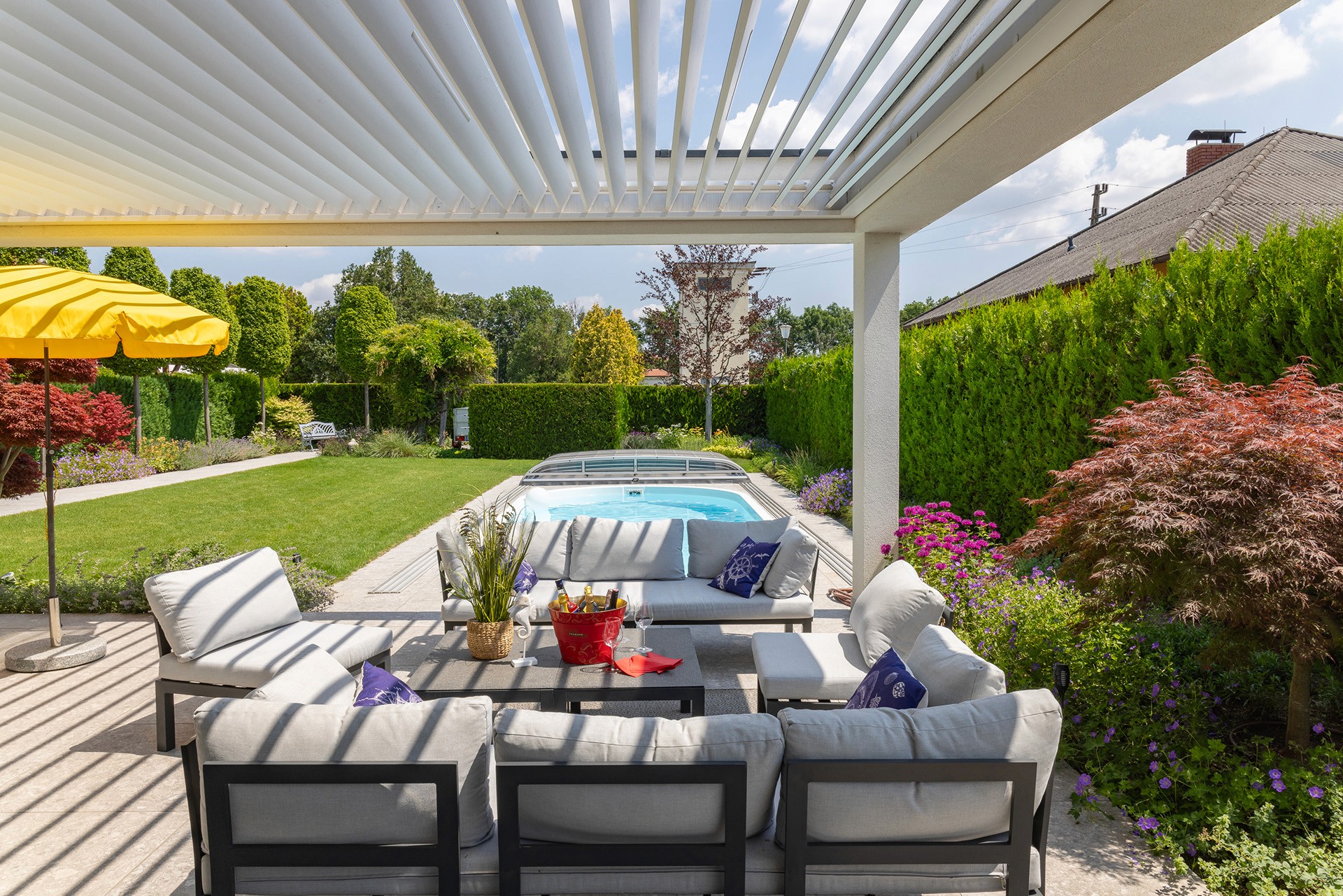 Überdachte Terrasse mit Sitzgruppe und Blick auf Pool, Rasenfläche und strukturierten Garten.