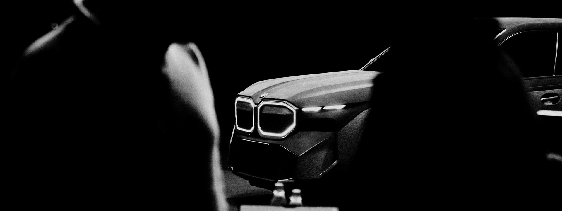 Black-and-white image of the front of a BMW XM framed by two people in the foreground