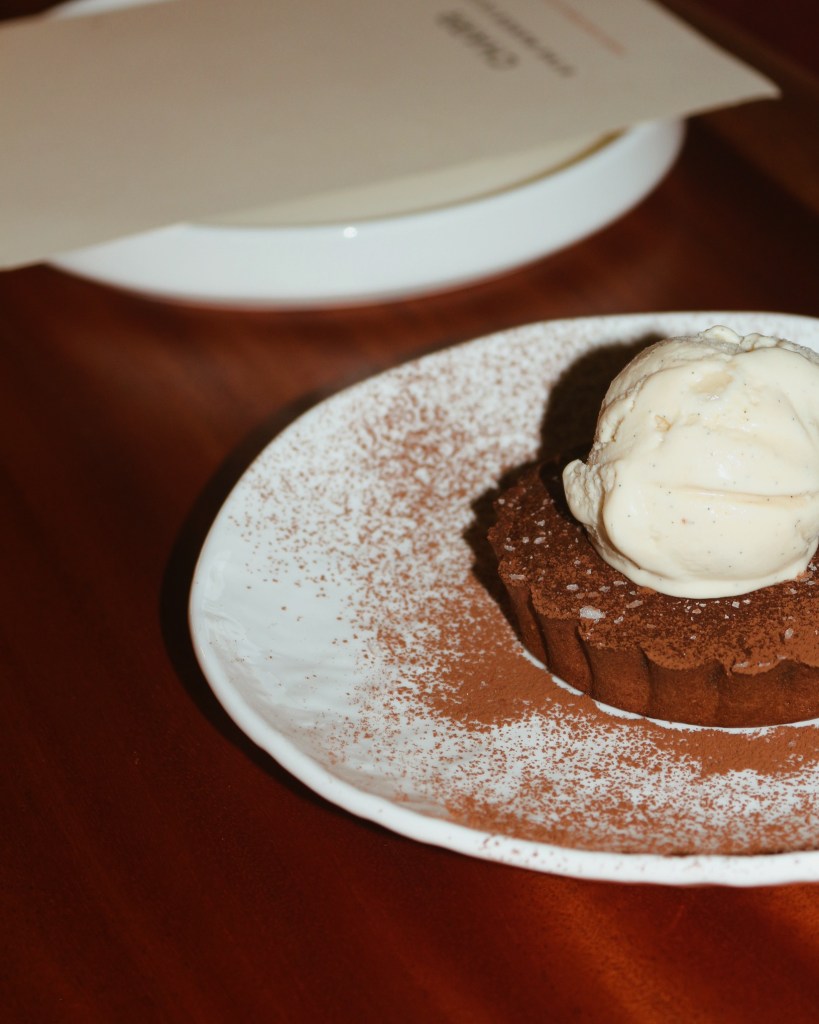 A chocolate dessert tart dusted with cocoa powder, topped with a scoop of vanilla ice cream, served on a white plate against a wooden table background.