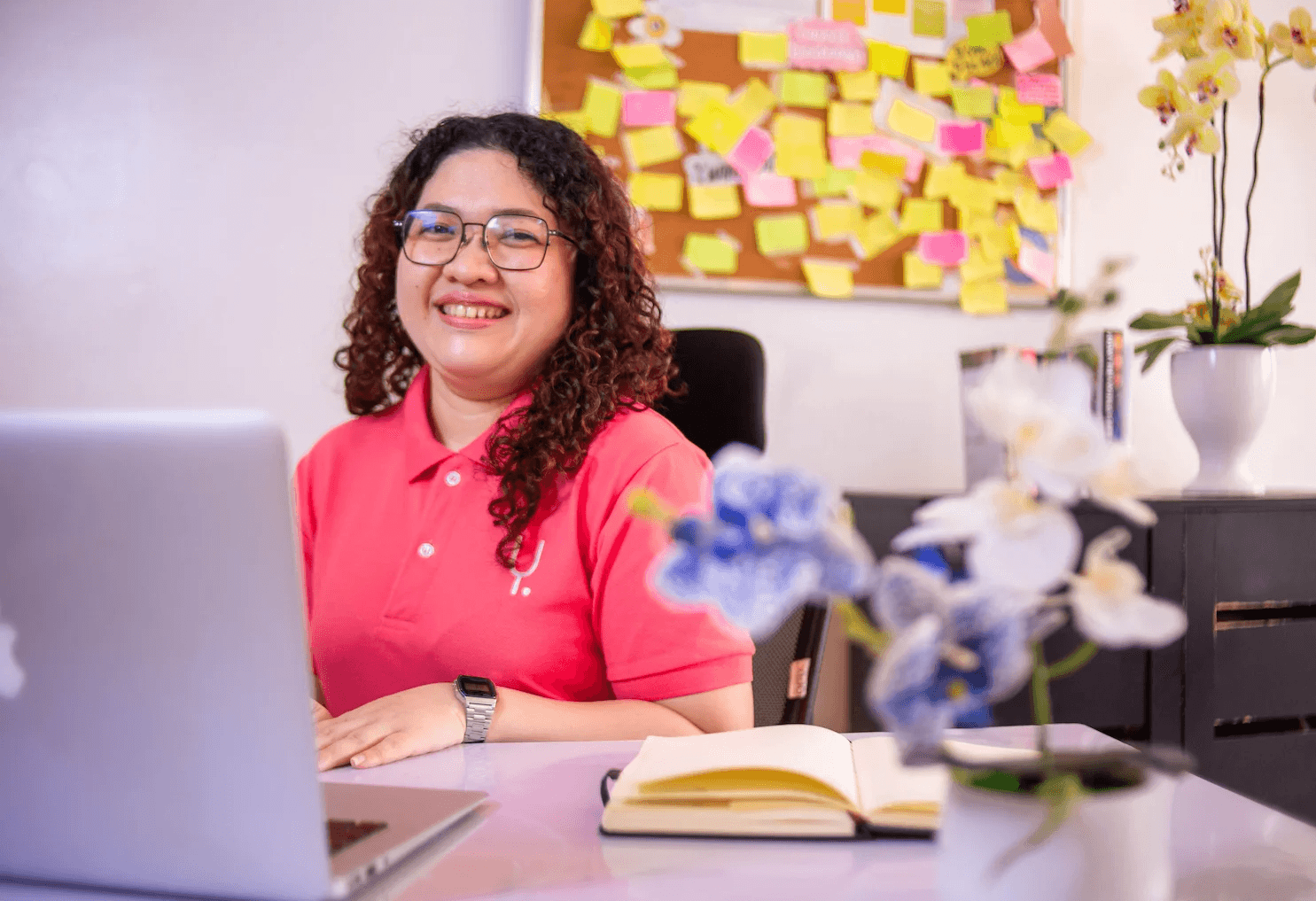 A smiling woman sits at a desk with a laptop and open notebook, with a colorful sticky note board and orchids in the background.