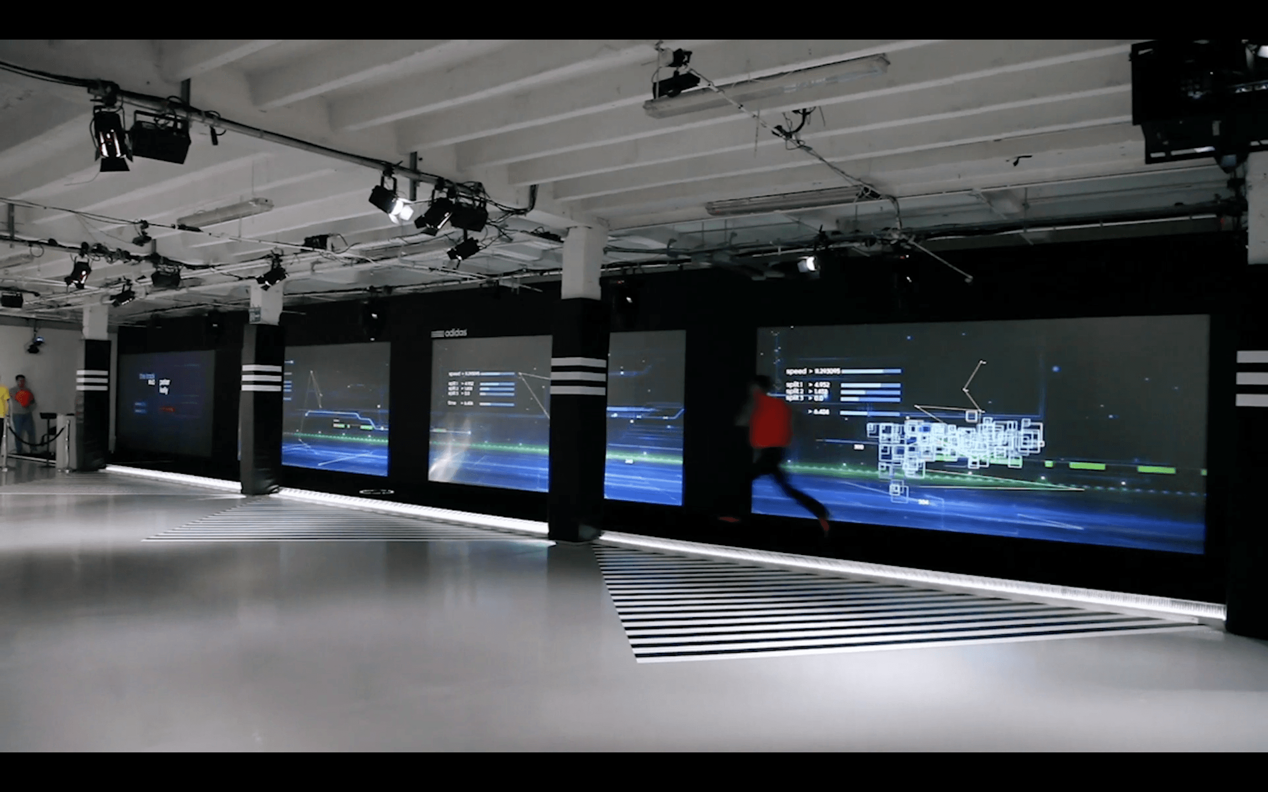 An individual runs across a spacious, modern indoor space featuring large digital screens displaying dynamic blueprints and data visualizations, under an array of spotlights.