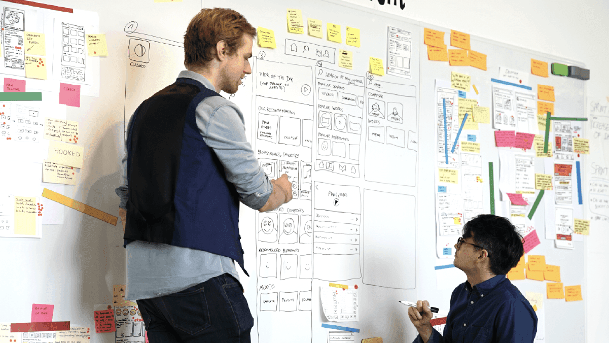 Two UX designers collaborating on wireframes and sticky notes on a whiteboard wall