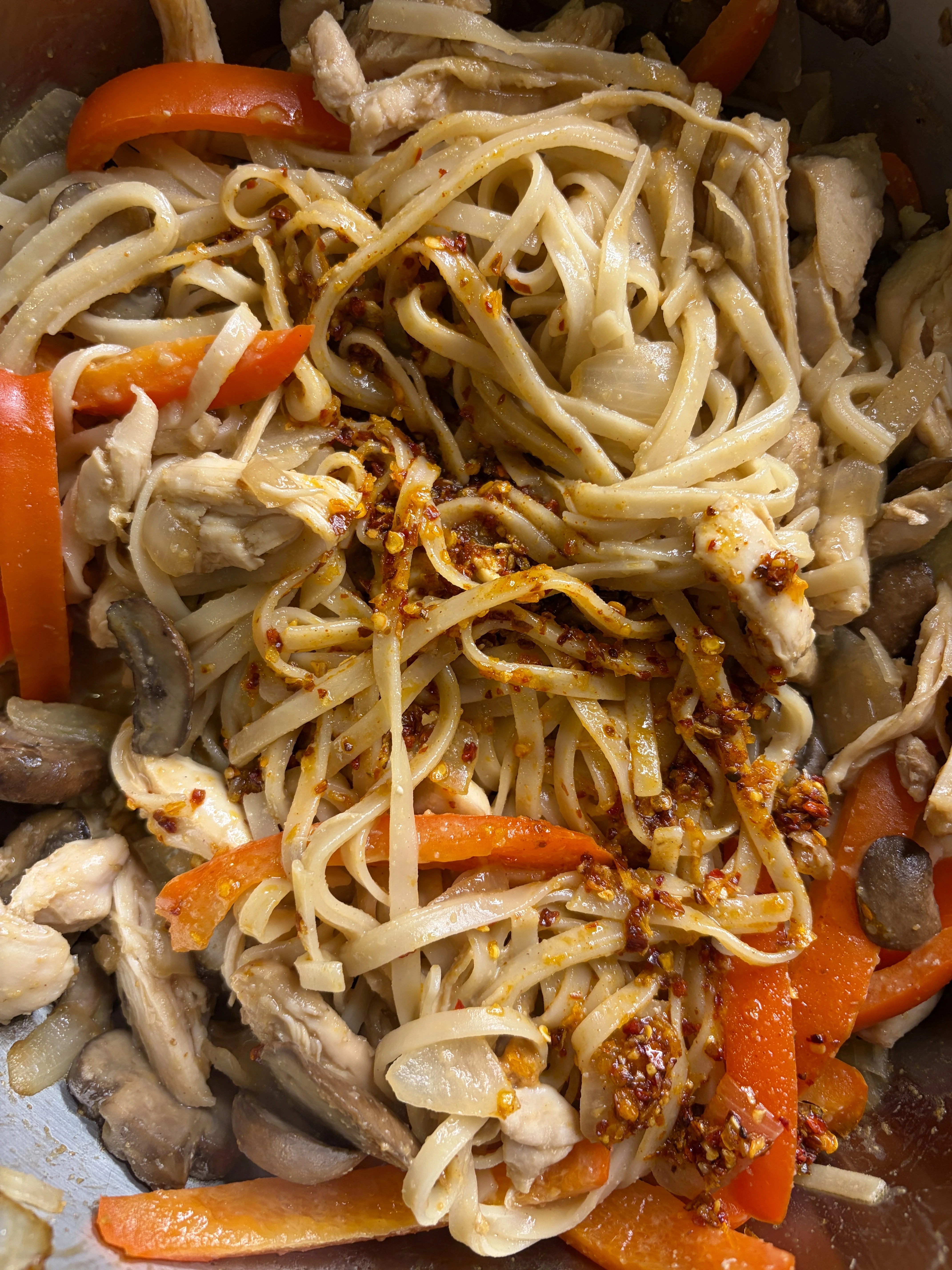 A bowl of gluten-free chicken peanut pad Thai made with sautéed chicken thighs, red bell pepper, mushrooms, and onion tossed with brown rice noodles in a creamy peanut-coconut amino sauce, then topped with crunchy chili oil, fresh cilantro, and a squeeze of lime.