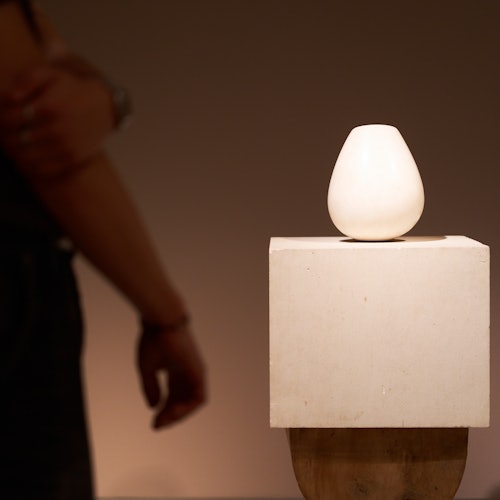 Eine kleine, weiße Vase steht auf einem quadratischen Sockel. Eine Person steht daneben, die Arme verschränkt, in einem schwach beleuchteten Raum.