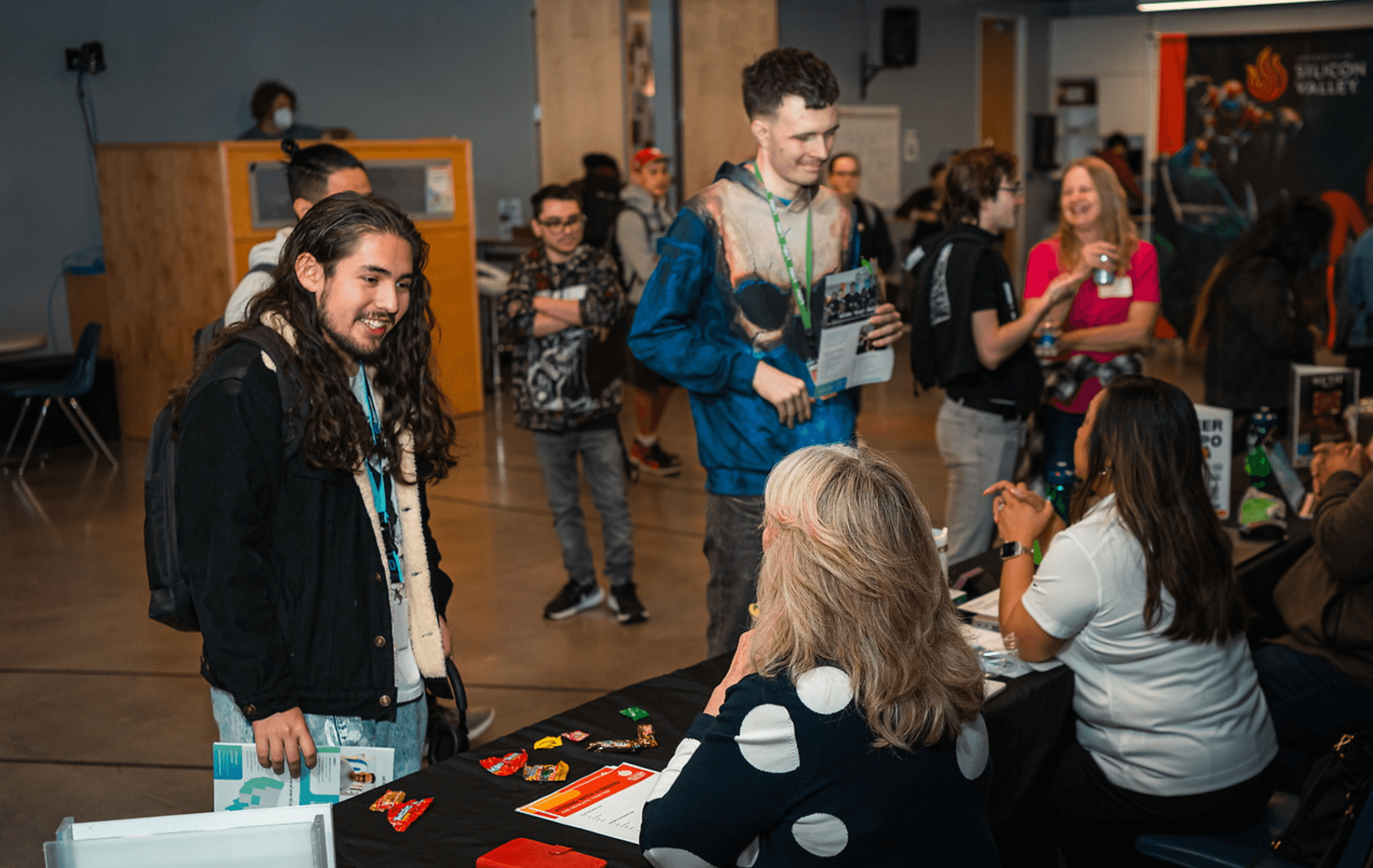 At a bustling job fair, a diverse group of young adults engages in conversations with recruiters seated at a table adorned with flyers and colorful candy, set against a backdrop of informational posters and a lively crowd, emphasizing networking and career opportunities.