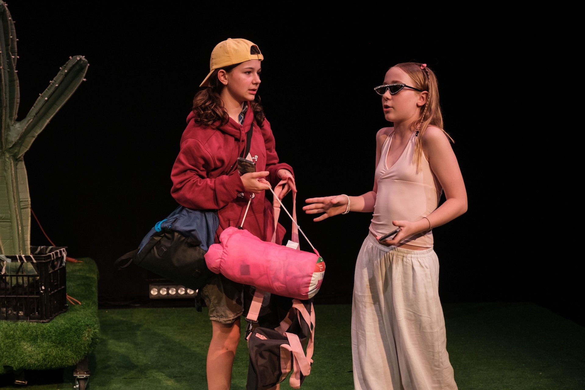 Two young performers stand facing each other on a grass-covered stage. One wears a backwards cap and carries camping gear, while the other, dressed in white with rhinestone sunglasses, gestures as they talk.
