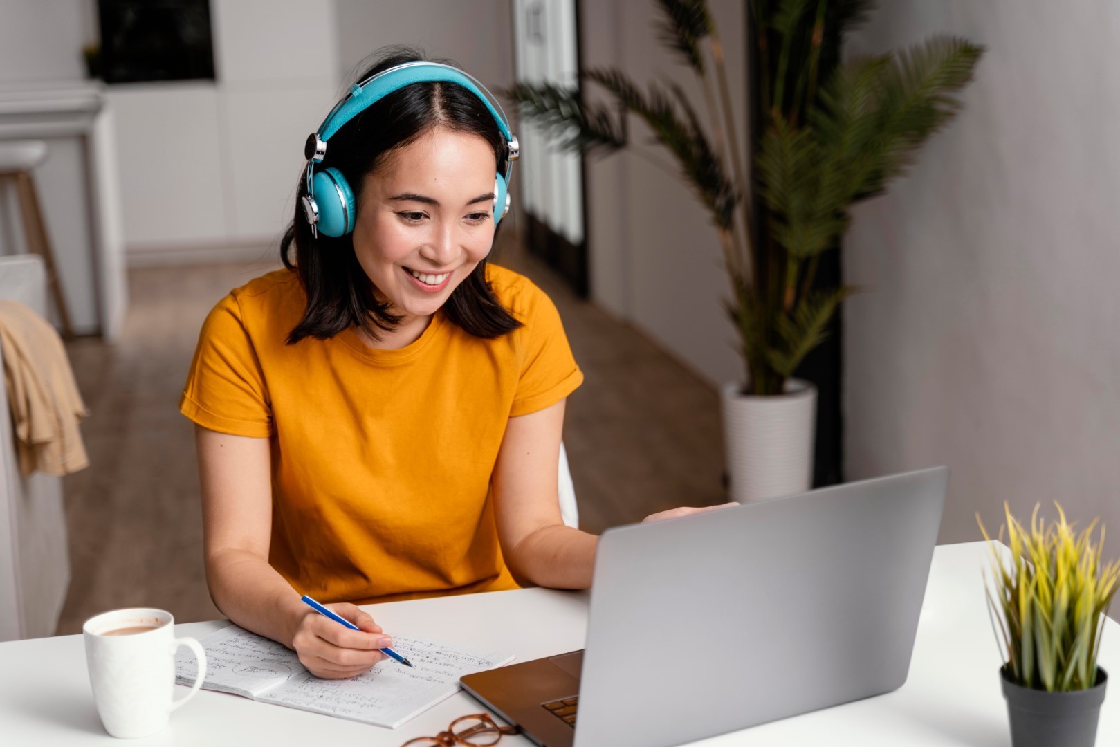 A woman wearing headphones and a yellow shirt smiles while working on a laptop at a modern desk, with a cup of coffee and potted plants nearby, illustrating a productive home office environment.