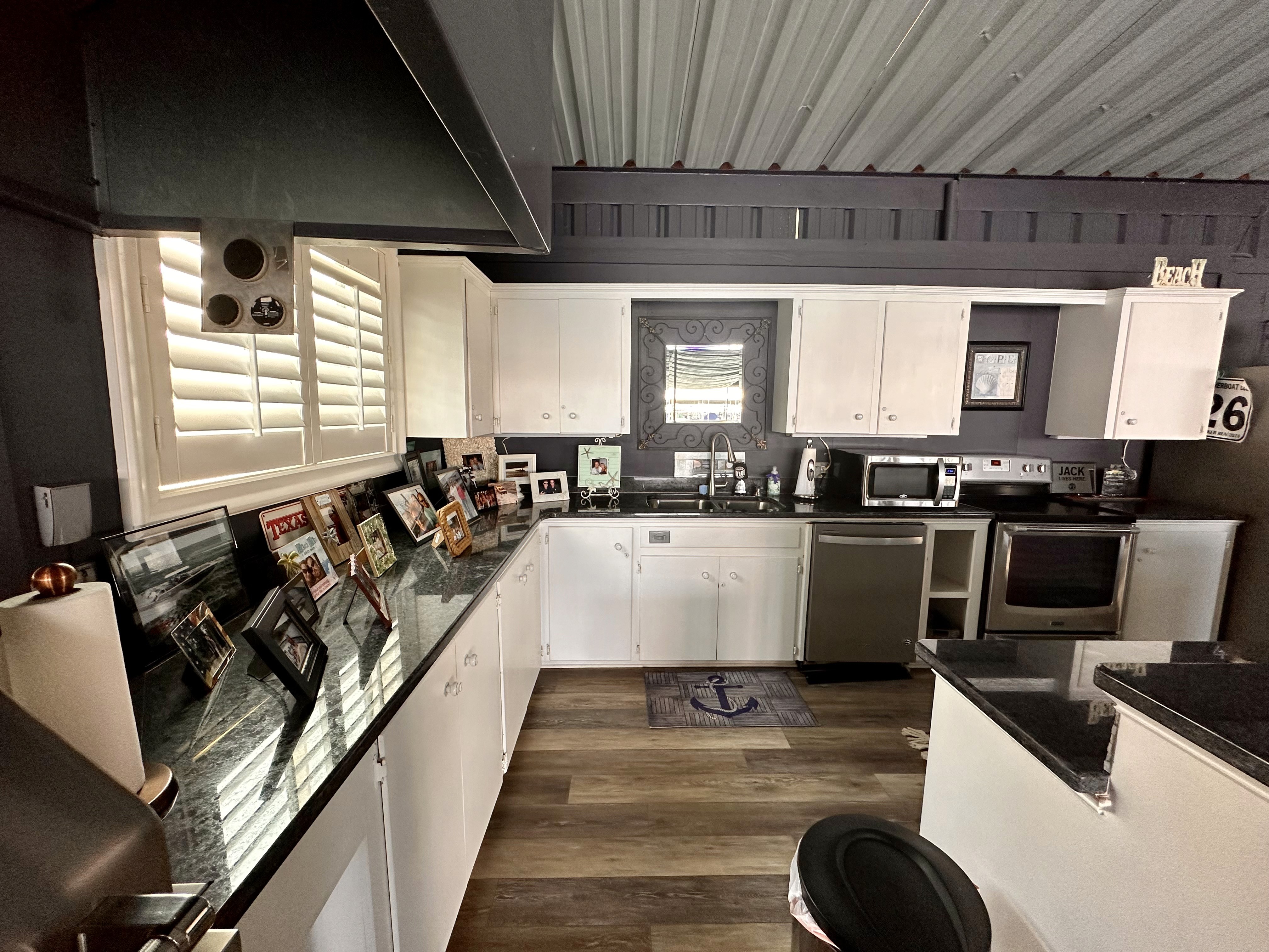 This modern kitchen features a sleek design with white cabinets, black granite countertops, stainless steel appliances, and a row of framed photos on display, illuminated by natural light from a window with shutters.