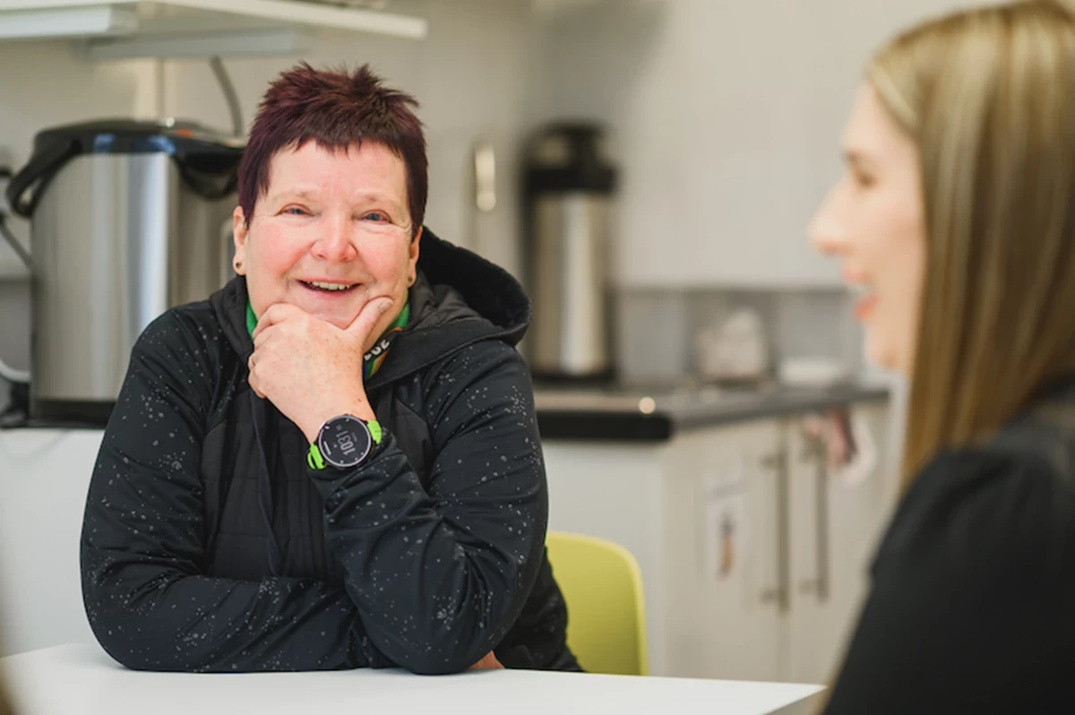 Two people sit across from each other at a table in a bright kitchen‑style room. One person rests their hand near their chin and wears a dark jacket and smartwatch.