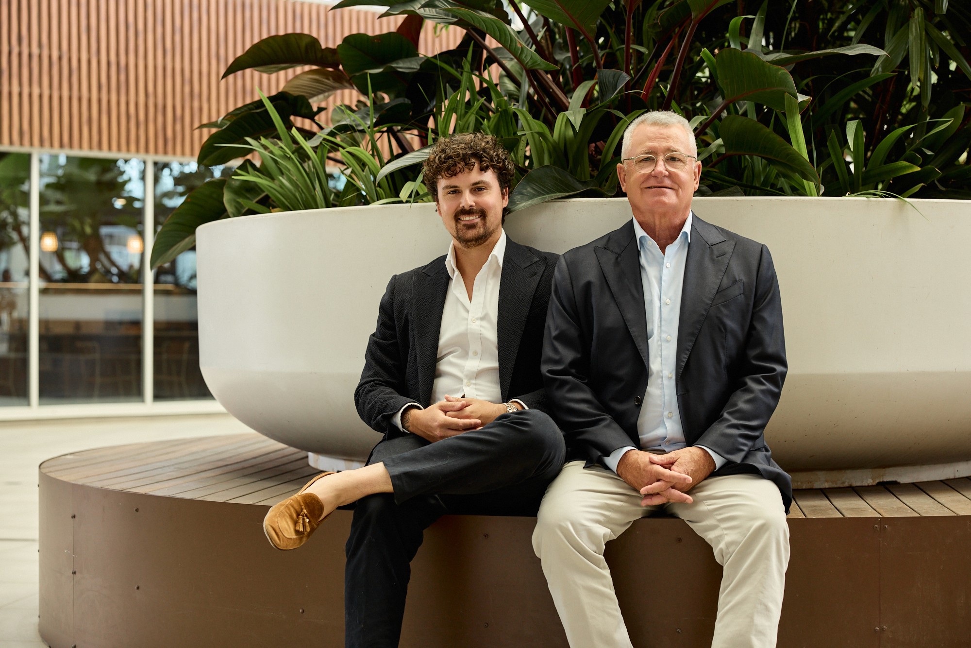 Three people collaborating in a modern office space behind glass with plants and warm lighting.