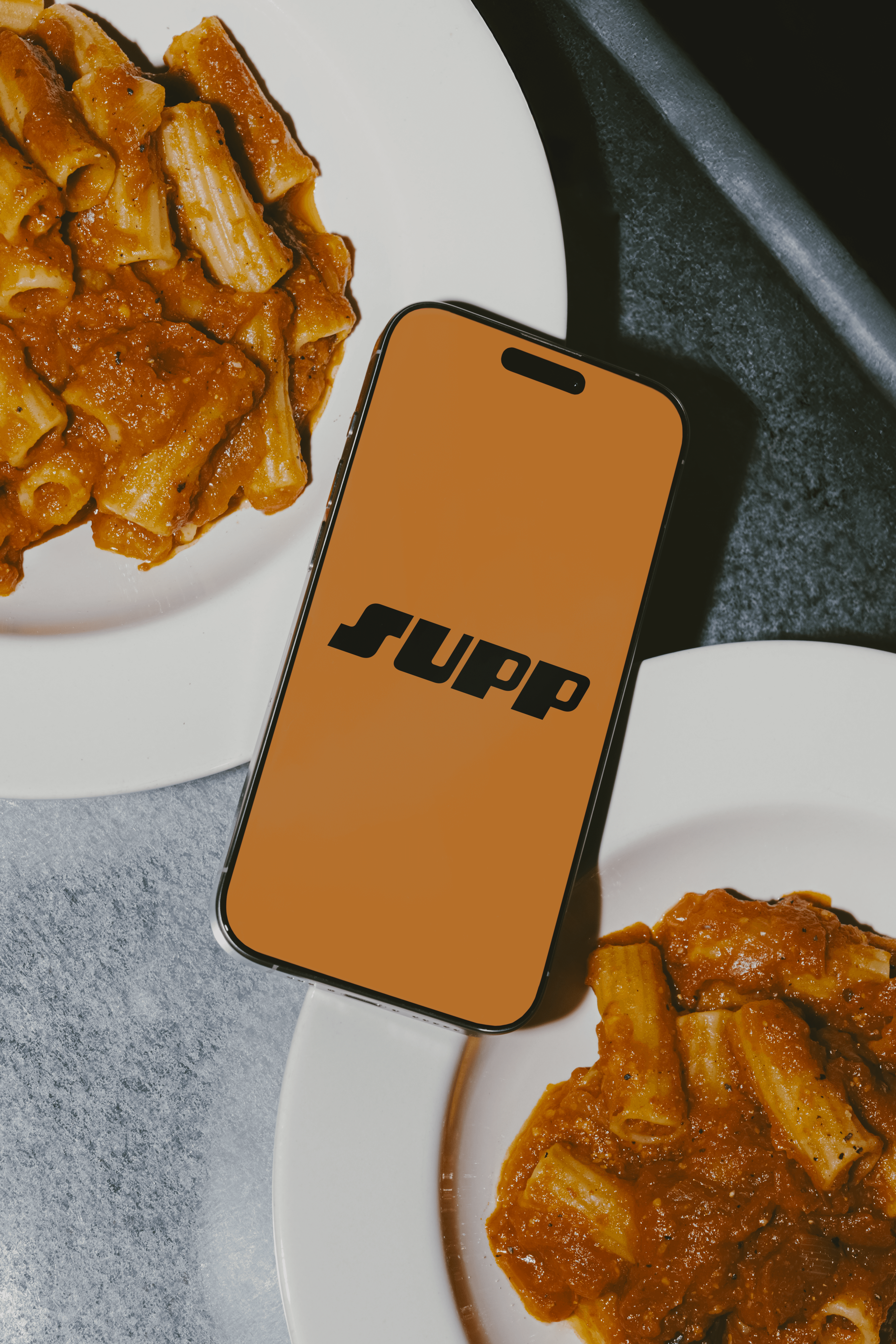 A phone displaying the Supp app logo, surrounded by plates of rigatoni