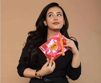 Woman holding colorful snack packets while looking upward against a beige background.