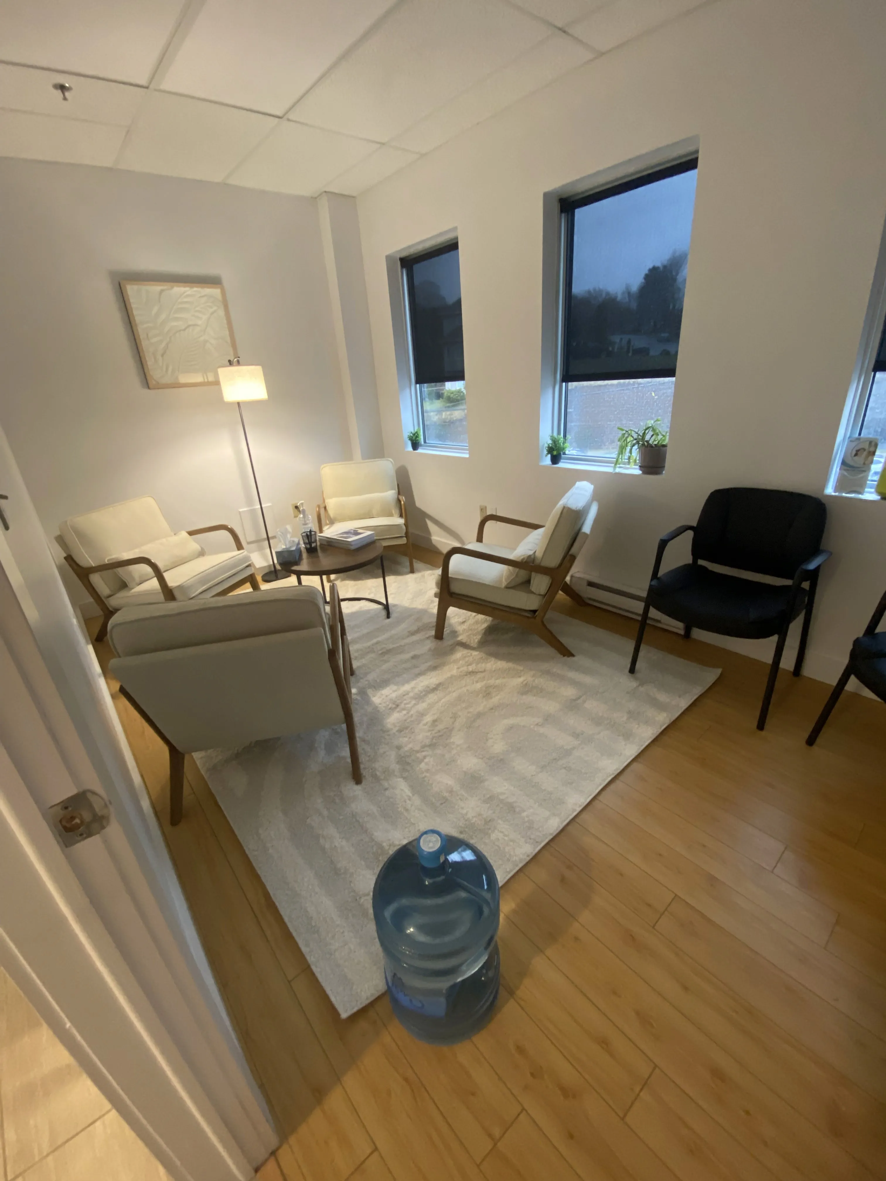 Waiting room with cream and gray armchairs, a black office chair, a floor lamp, and windows overlooking the street