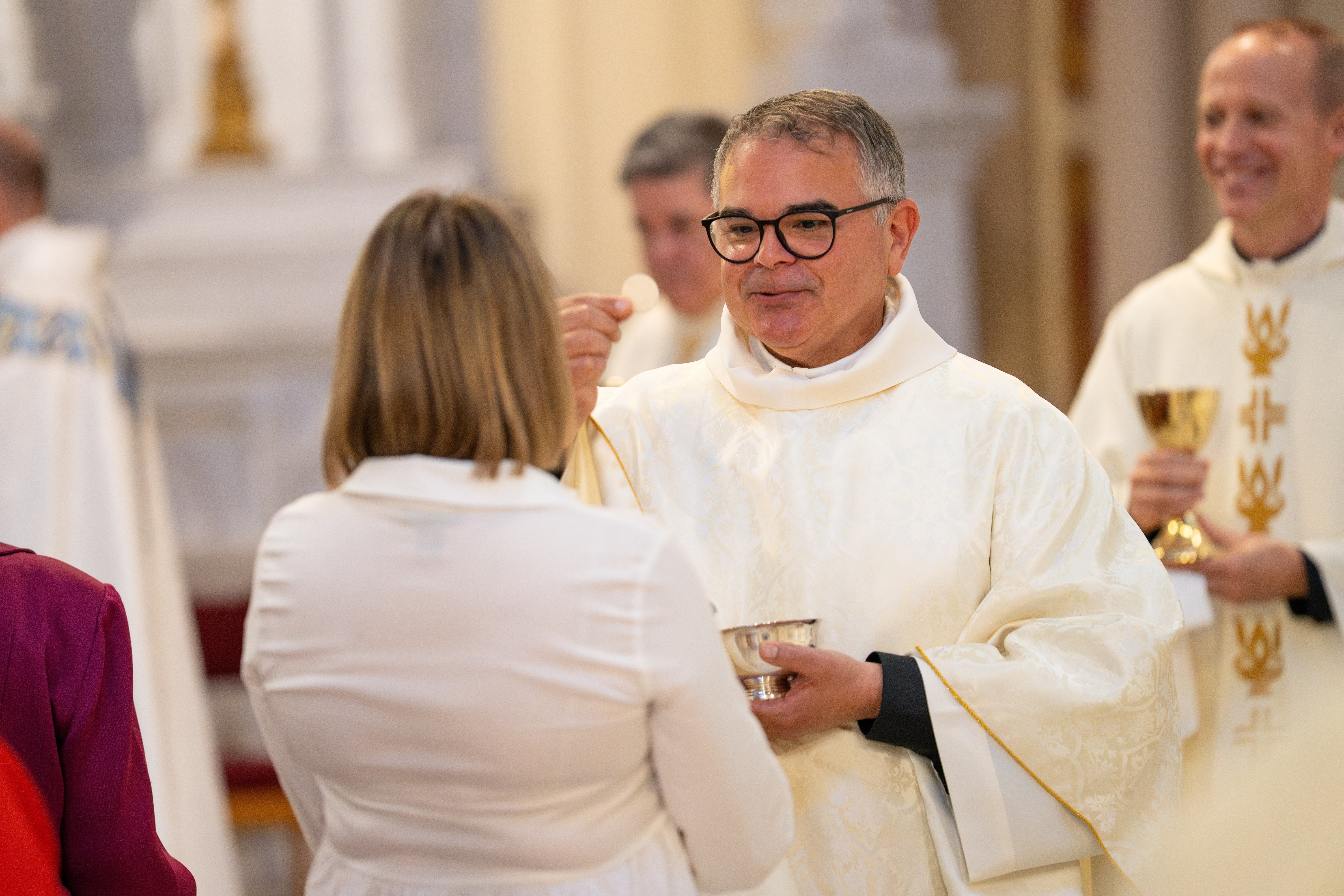 A priest wearing white robes interacts with a woman in a religious setting, surrounded by other clergy.