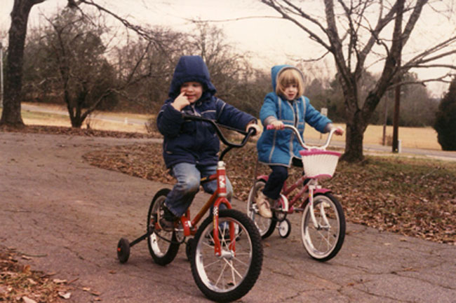 Two young children are riding bicycles on a paved path. The child on the left is on a red tricycle with a training wheel, wearing a dark blue hooded jacket and jeans. The child on the right is on a pink bicycle with a white basket, wearing a light blue hooded jacket and dark pants. The path is surrounded by leaf-covered ground and trees with bare branches, indicating it might be autumn or winter. The sky is overcast.