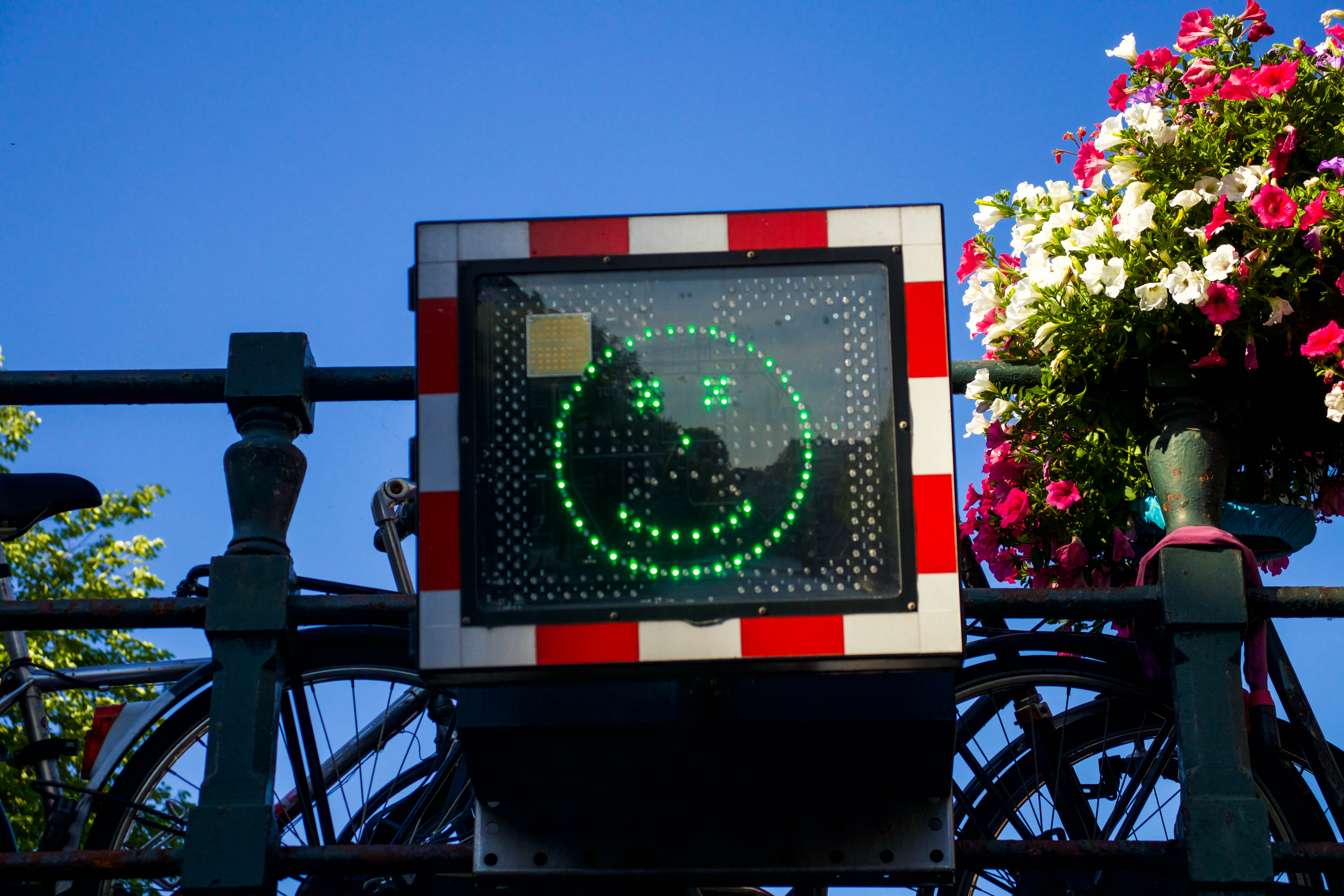 Smiling green bicycle traffic signal on a bridge with flowers and parked bikes in the background.